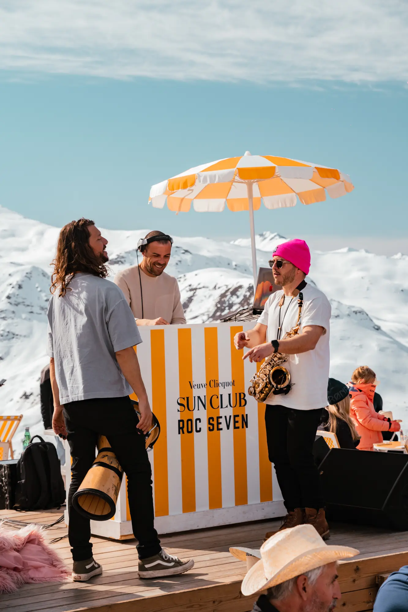 Trois hommes discutent sur une terrasse en bois avec un stand rayé jaune et blanc marqué Veuve Clicquot Sun Club Roc Seven, entouré de montagnes enneigées, l'un portant un saxophone et un bonnet rose, un autre tenant un grand tambour, et un DJ souriant derrière le stand sous un parasol rayé jaune et blanc.