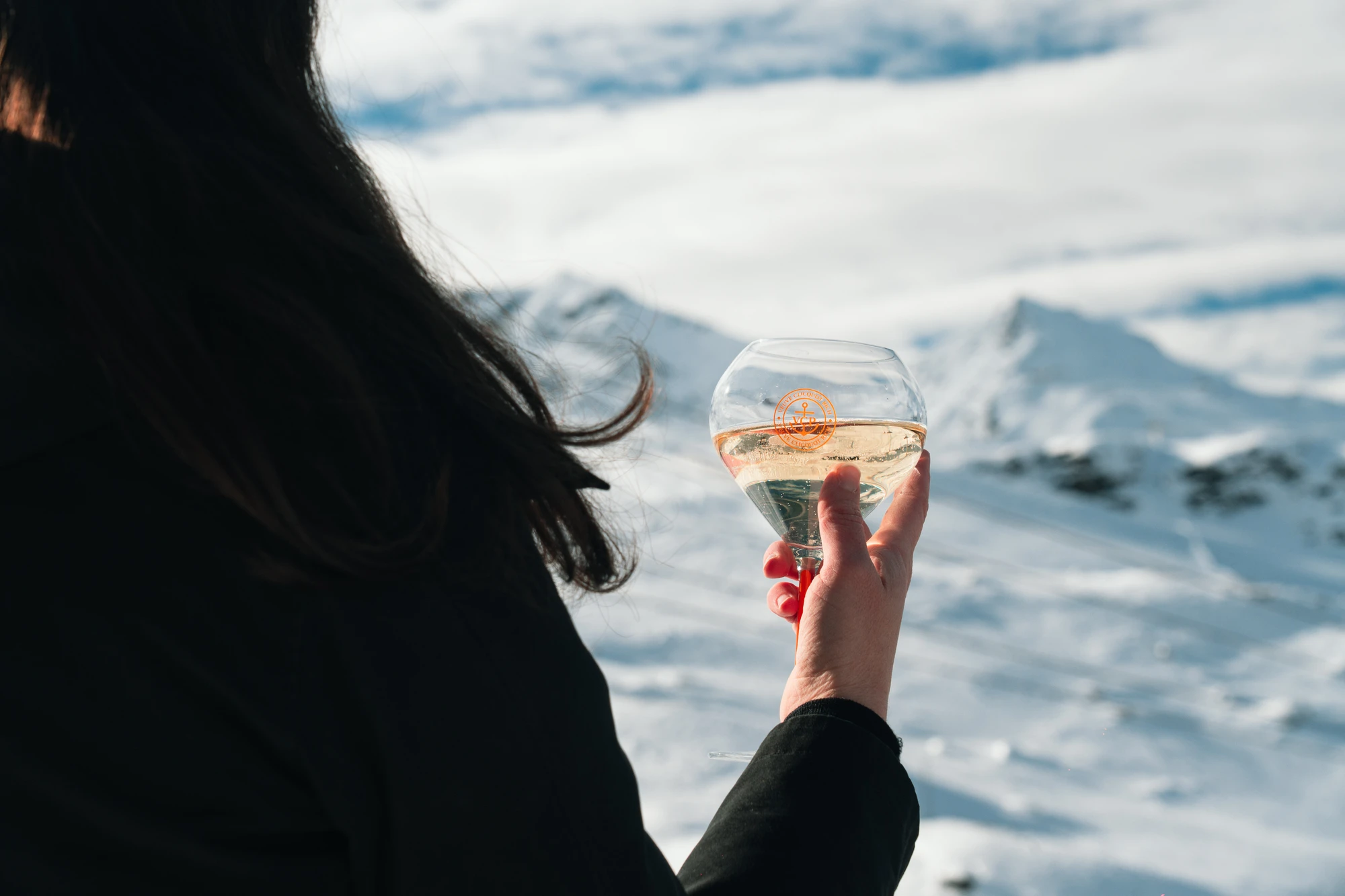 Person tenant un verre de champagne devant un paysage de montagnes enneigées sous un ciel partiellement nuageux.
