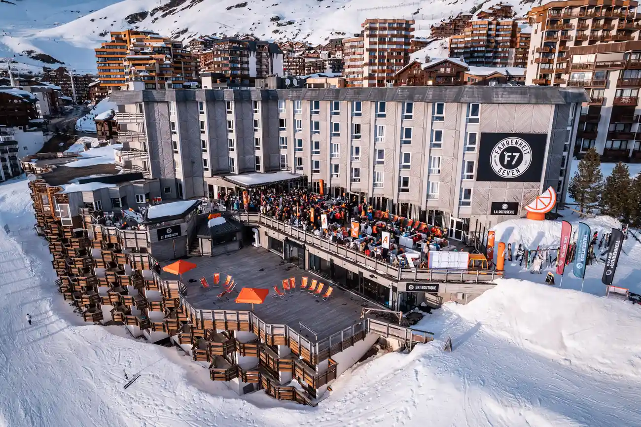 Grand bâtiment hôtelier moderne sur un domaine skiable enneigé avec une grande terrasse animée de personnes, chaises longues orange, parasols et bannières publicitaires, entouré de nombreuses résidences montagnardes en arrière-plan.