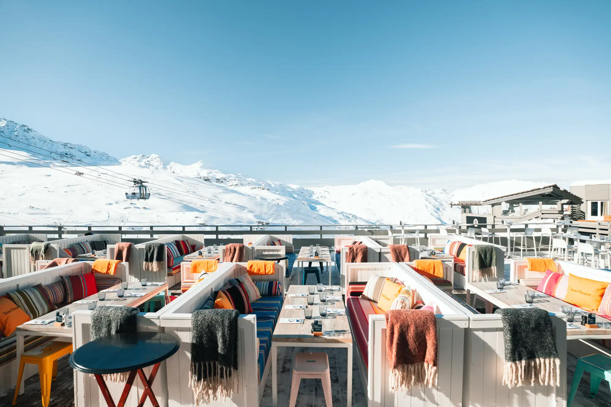 Terrasse d'un restaurant avec des banquettes blanches garnies de couvertures colorées et coussins, surplombant un paysage de montagnes enneigées sous un ciel bleu clair, avec une cabine de téléphérique visible à gauche.