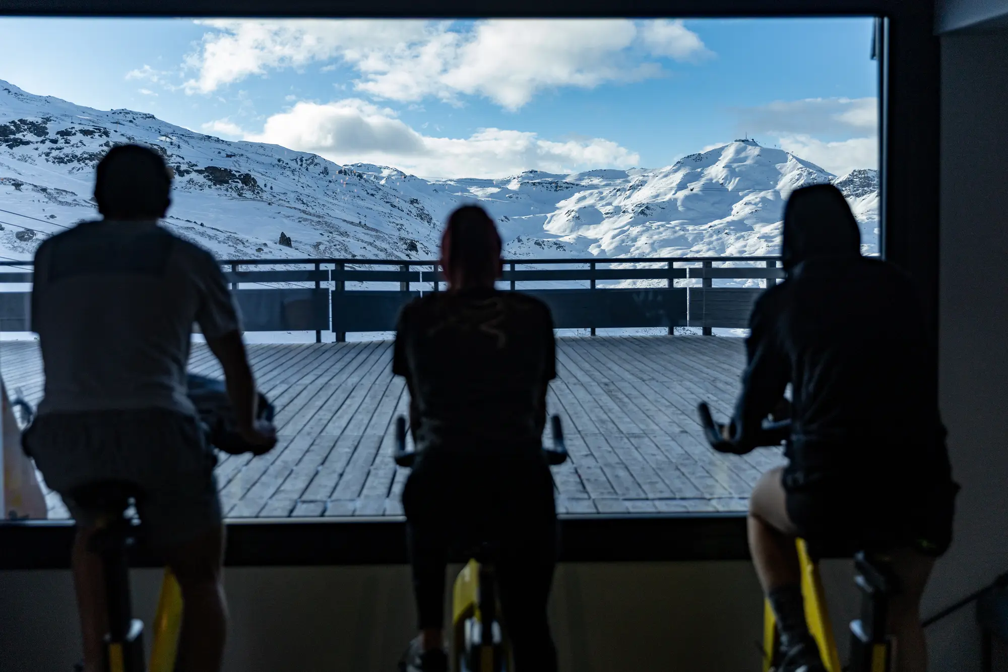 Trois personnes à vélo stationnaire en intérieur regardant par une grande fenêtre vers une terrasse en bois et des montagnes enneigées sous un ciel bleu avec quelques nuages.