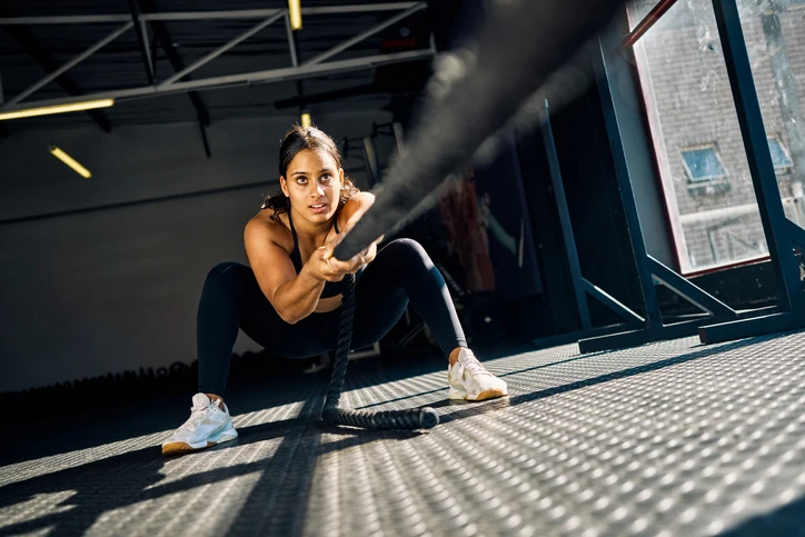 Femme effectuant un exercice de corde ondulatoire dans une salle de sport, accroupie avec une expression concentrée.