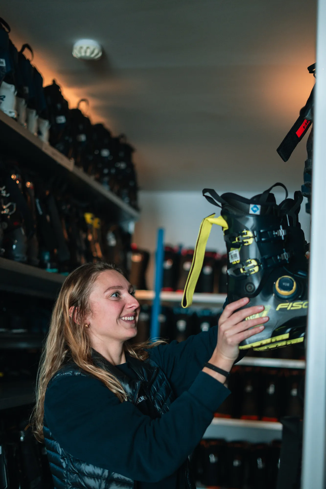 Une femme souriante choisit une chaussure de ski noire avec des boucles jaunes dans un local de rangement rempli de chaussures de ski alignées sur des étagères.