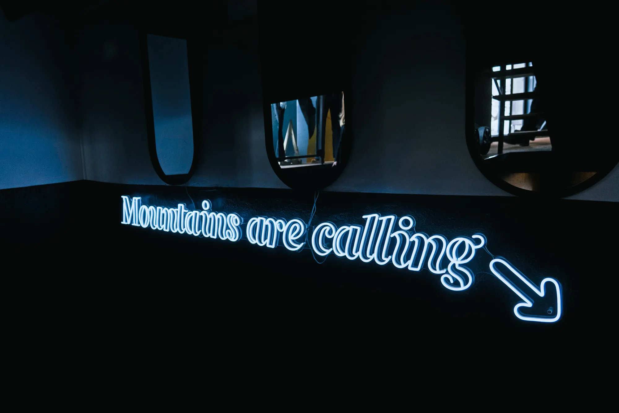 Enseigne lumineuse bleue avec le texte « Mountains are calling » et une flèche pointant vers la droite sur un mur sombre sous trois petits miroirs noirs.