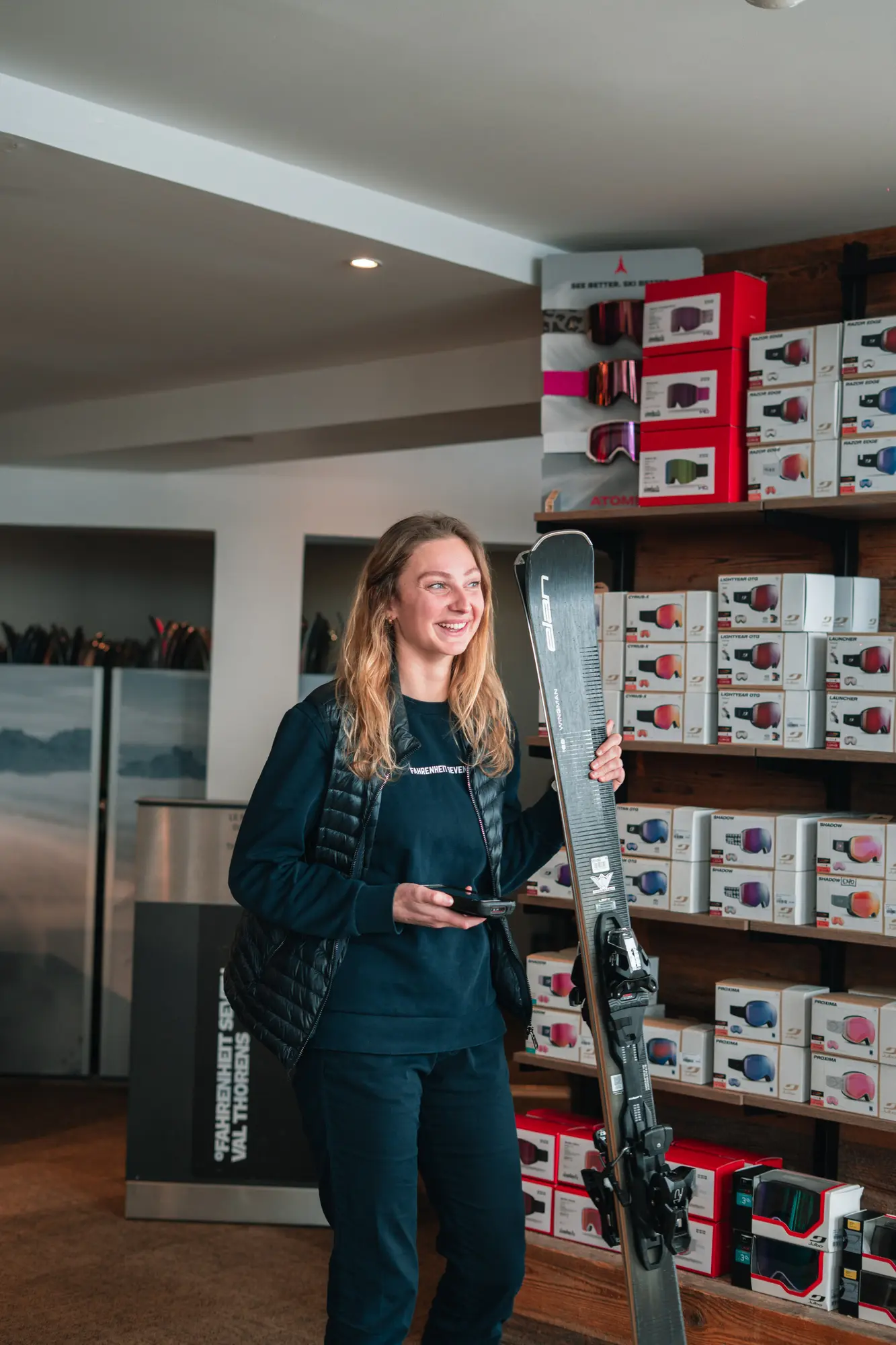 Jeune femme souriante tenant un ski gris Atomic dans une main et un appareil dans l'autre, debout dans un magasin de sports avec des étagères remplies de boîtes de lunettes de ski.