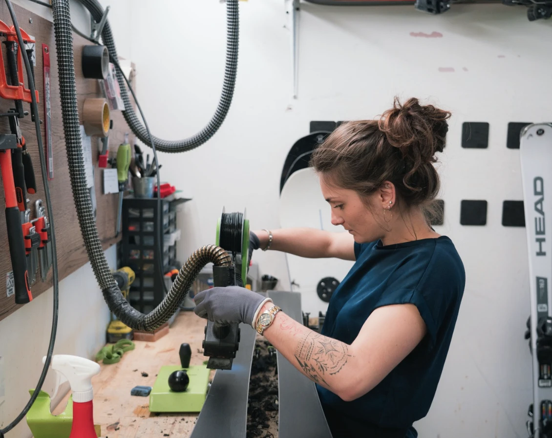 Une femme dans un atelier ajuste un équipement sur une paire de skis, portant un gant gris et une montre, avec plusieurs outils suspendus au mur à gauche.