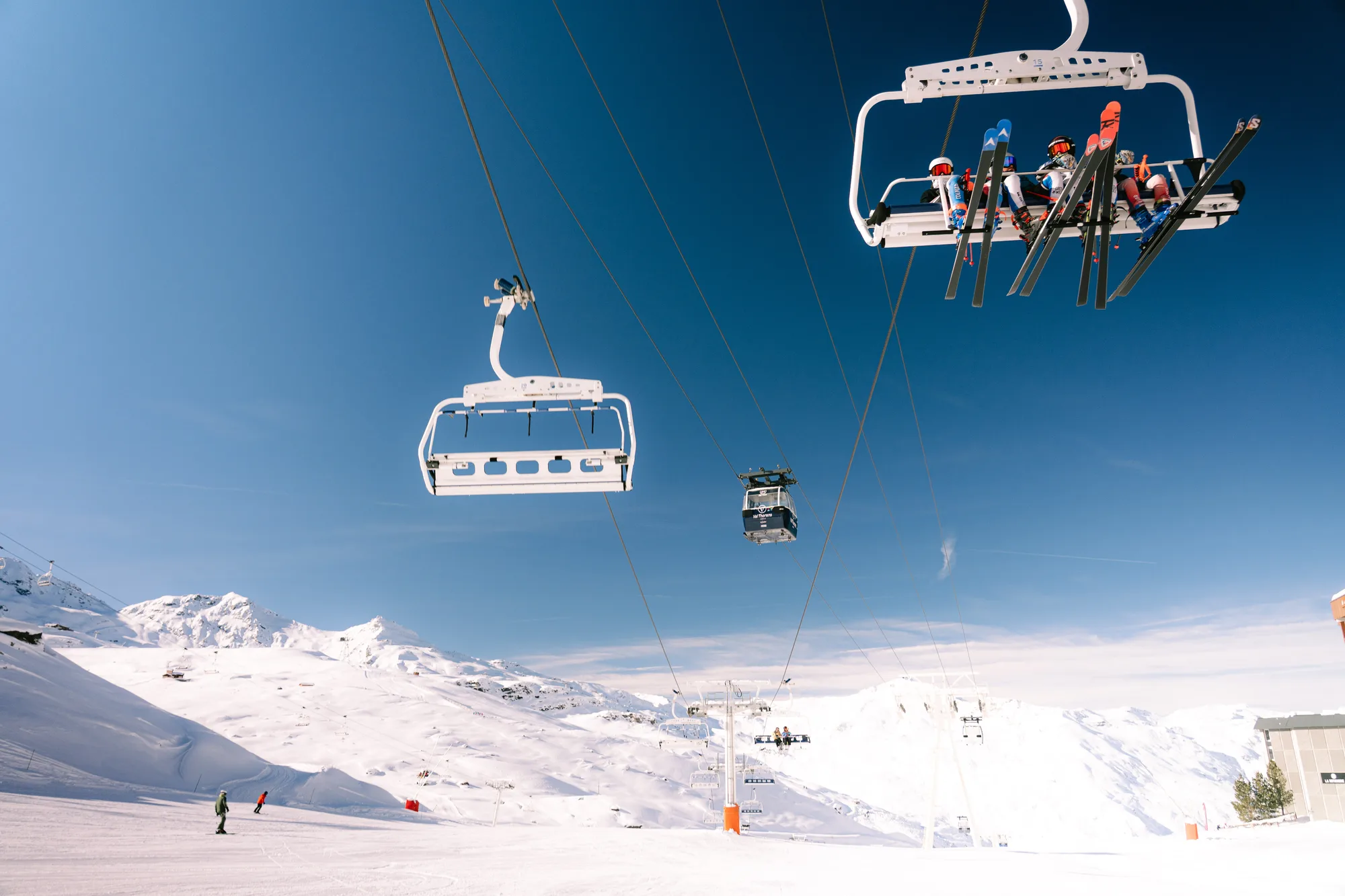 Télésiège blanc en montagnes enneigées avec plusieurs skieurs assis portant des vêtements de ski colorés, sous un ciel bleu clair, avec des skieurs descendant la pente en dessous.