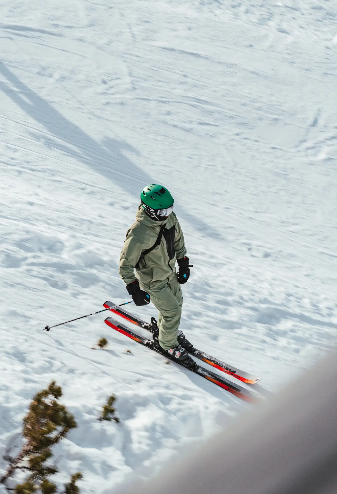 Un skieur portant un casque vert et des vêtements de ski beige descend une pente enneigée avec des skis noirs et rouges, tenant des bâtons de ski dans ses mains.