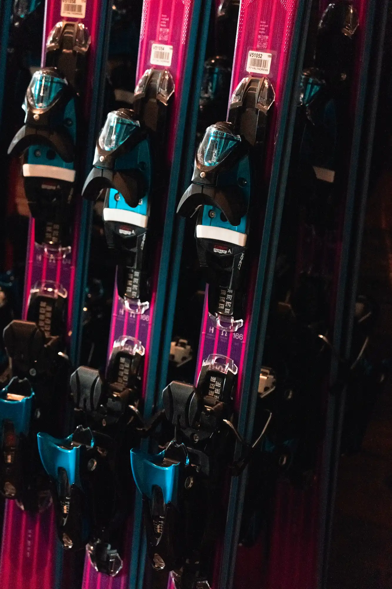 Plusieurs paires de skis roses avec des fixations noires et bleues alignées verticalement, suspendues dans un espace sombre.