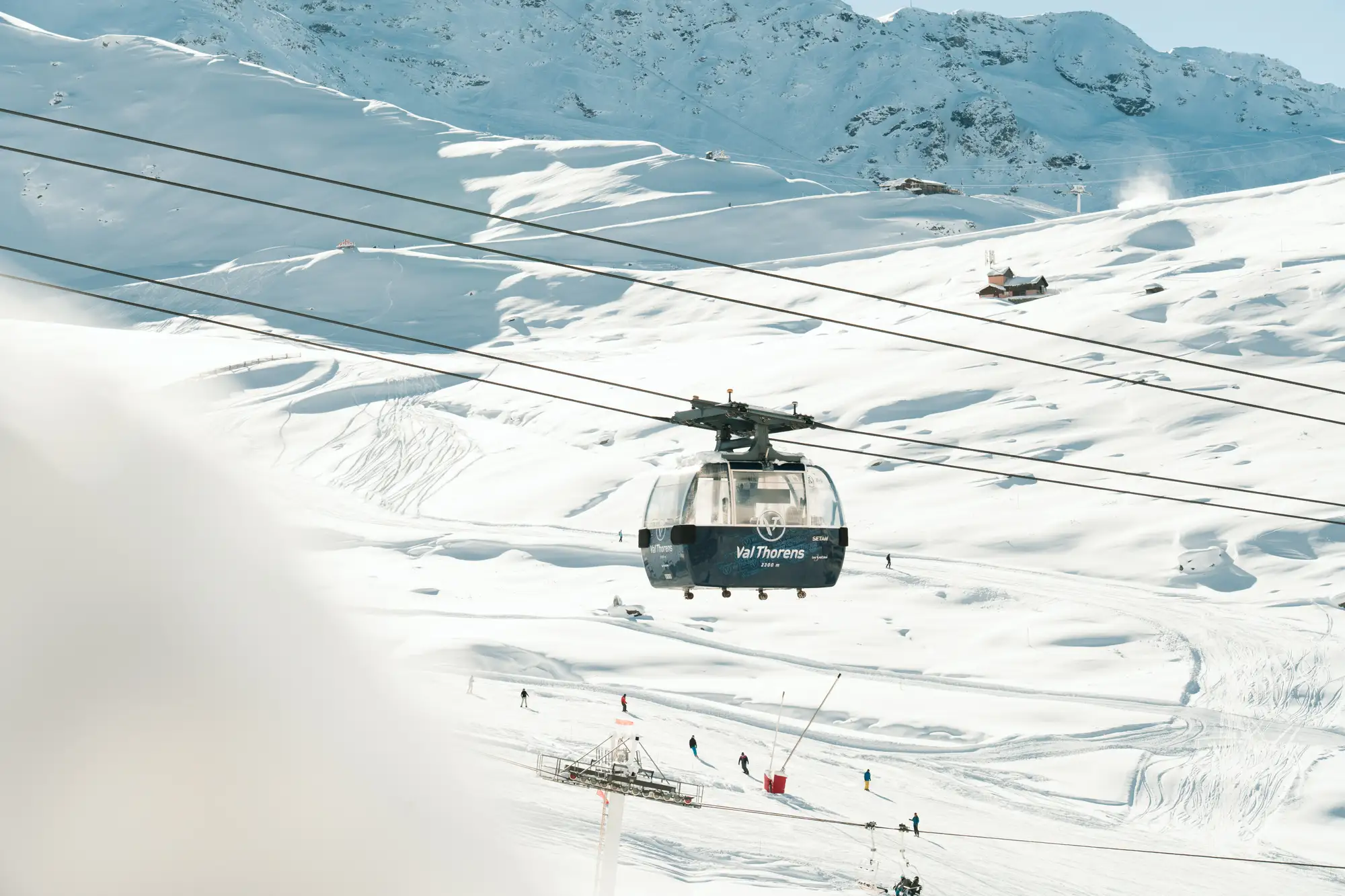 Téléphérique bleu portant l'inscription Val Thorens suspendu au-dessus d'une station de ski enneigée avec des skieurs visibles en dessous et des montagnes en arrière-plan.