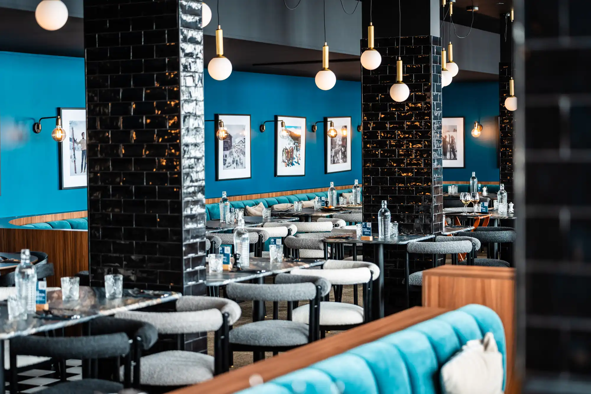 Intérieur moderne d'un restaurant avec murs bleus, colonnes carrelées noires, tables dressées de bouteilles d'eau et verres, chaises grises, et banquettes bleues, éclairé par des globes suspendus blancs.
