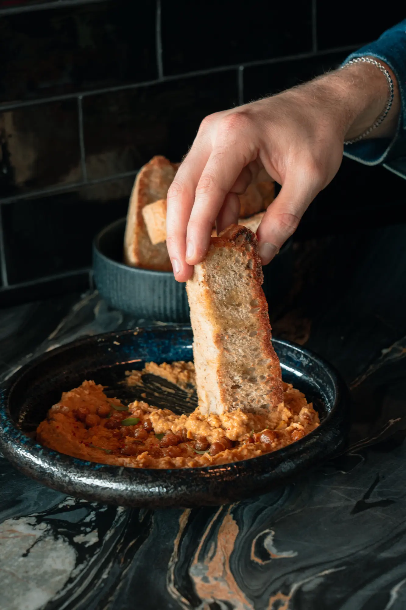 Une main plonge une tranche de pain grillé dans une purée de pois chiches épicée servie dans un bol noir sur une surface en marbre foncé.