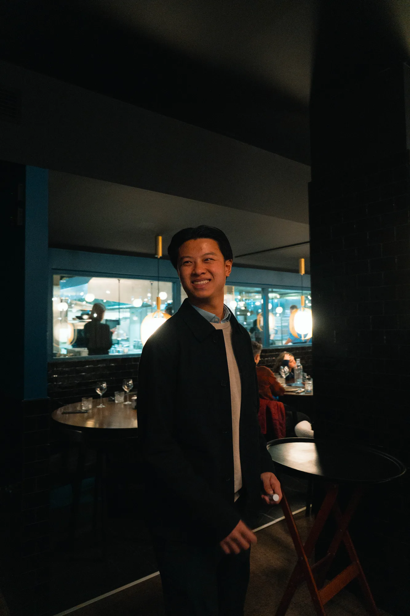Homme souriant debout dans un restaurant sombre, avec une table ronde en arrière-plan et des personnes assises au fond.
