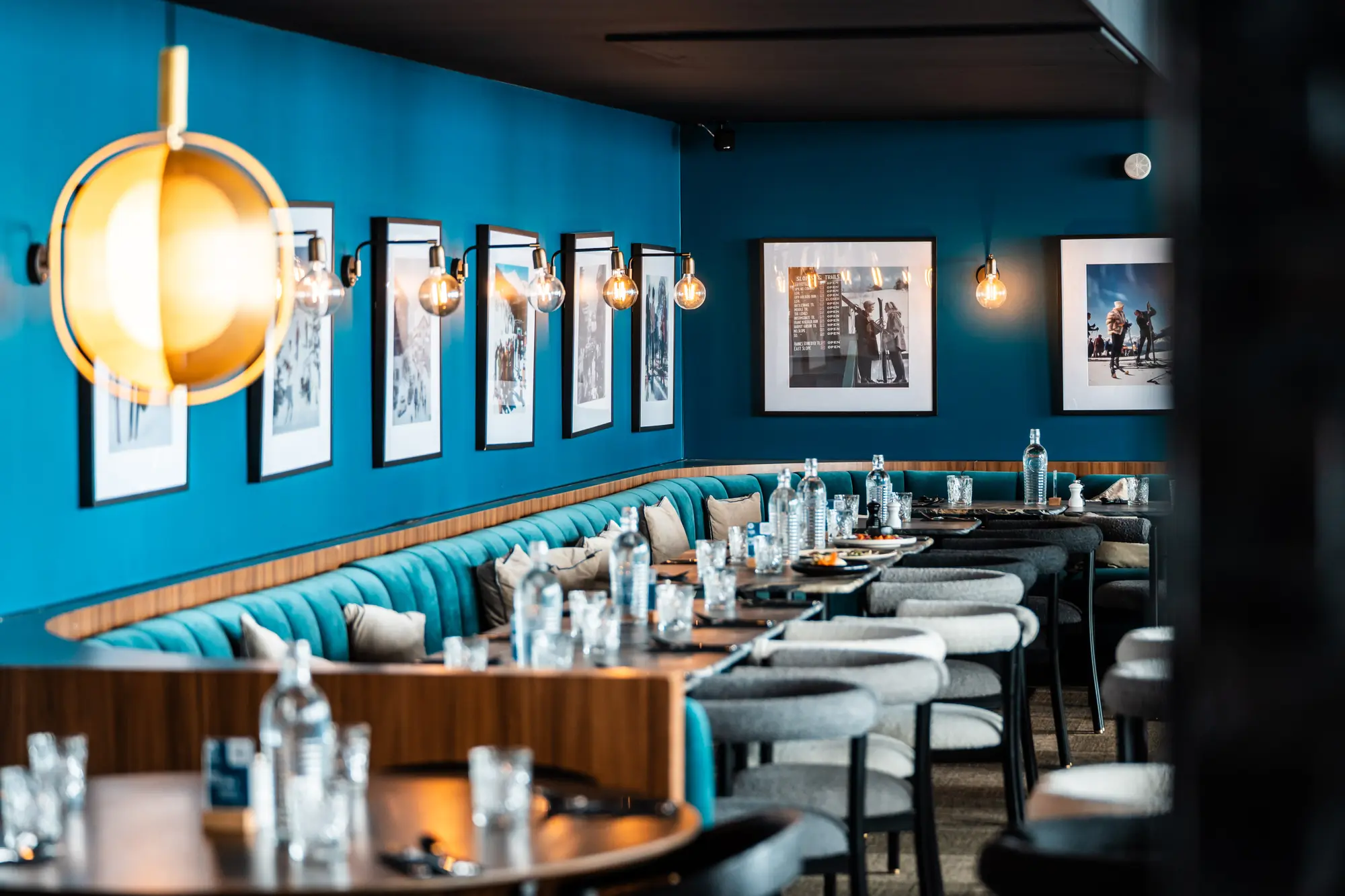 Intérieur d'un restaurant avec murs bleus décorés de photos en noir et blanc, banquettes bleu canard, tables en bois dressées avec bouteilles d'eau, verres et assiettes et chaises grises autour.