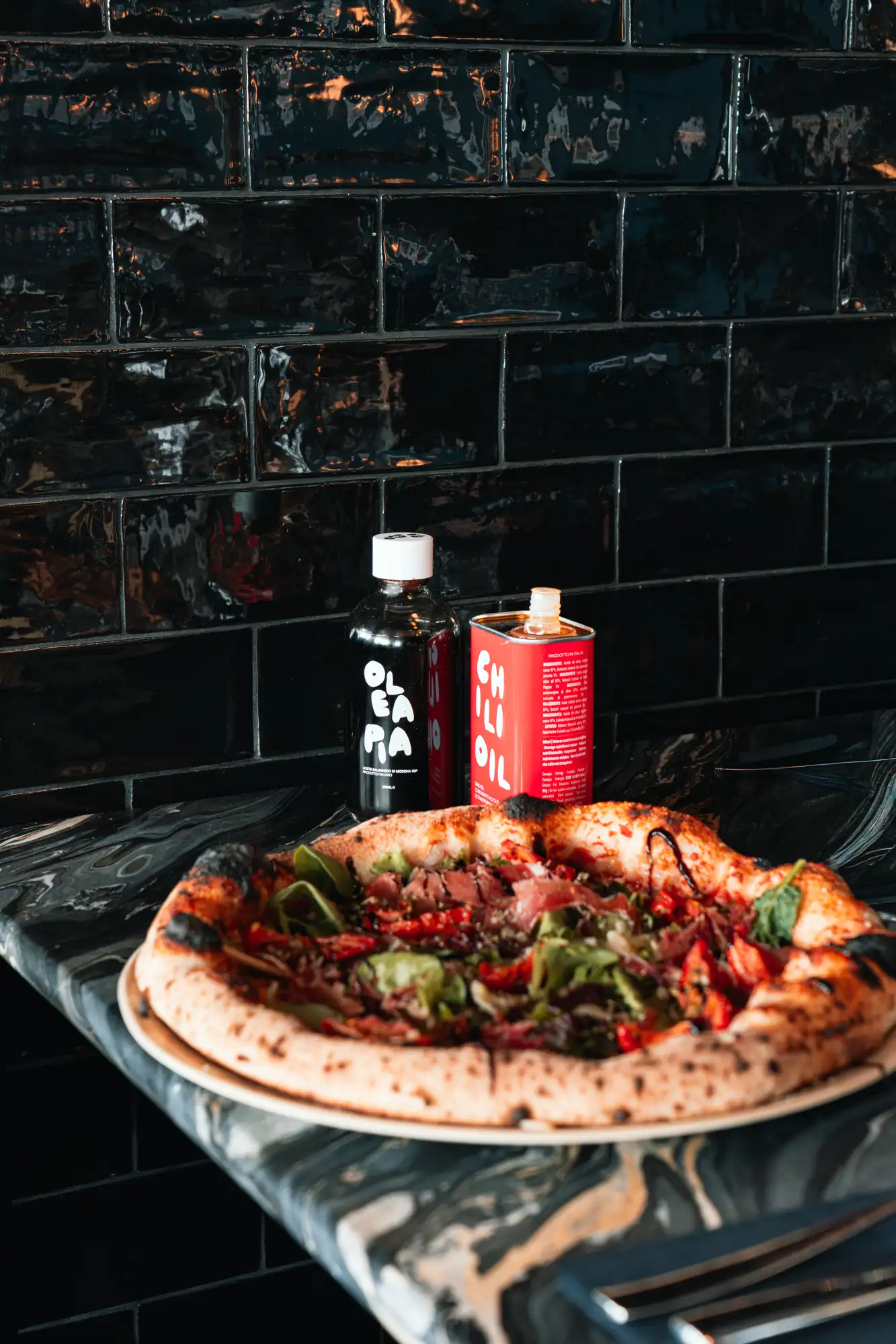 Une pizza garnie de légumes et viande, posée sur un comptoir en pierre noire, avec deux bouteilles contenant de l'huile d'olive et du vinaigre balsamique à l'arrière-plan devant un mur de carreaux noirs brillants.
