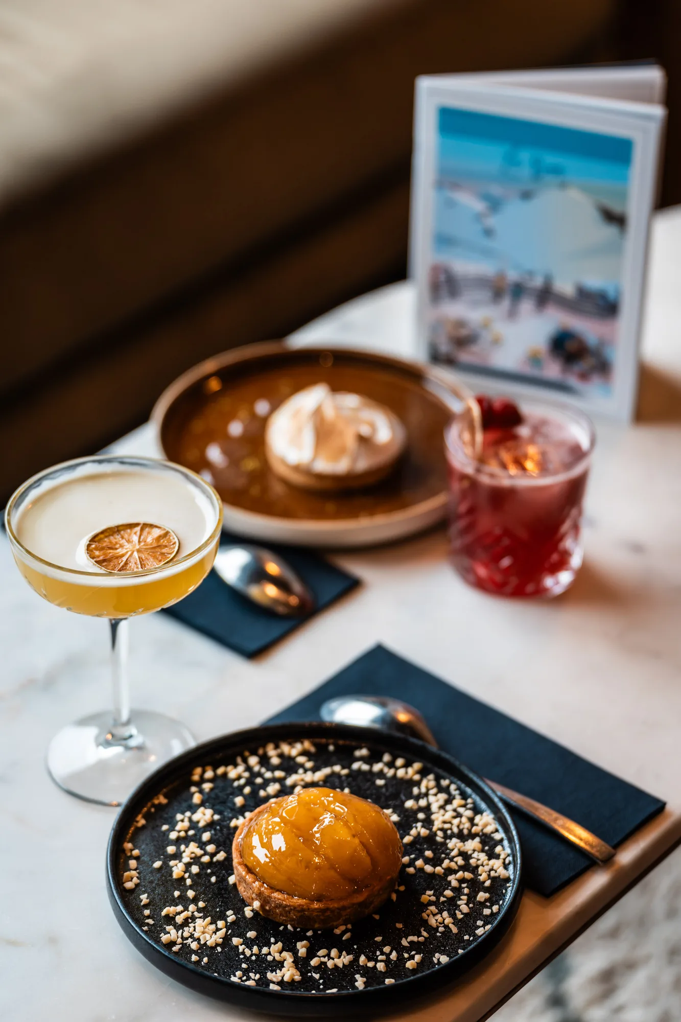 Table with two desserts on black plates, one topped with a glossy yellow fruit glaze and sprinkled with chopped nuts, the other with a dollop of cream, accompanied by two cocktails—one yellow with a dried citrus slice and the other red with a garnish—on a white marble surface with black napkins and spoons.