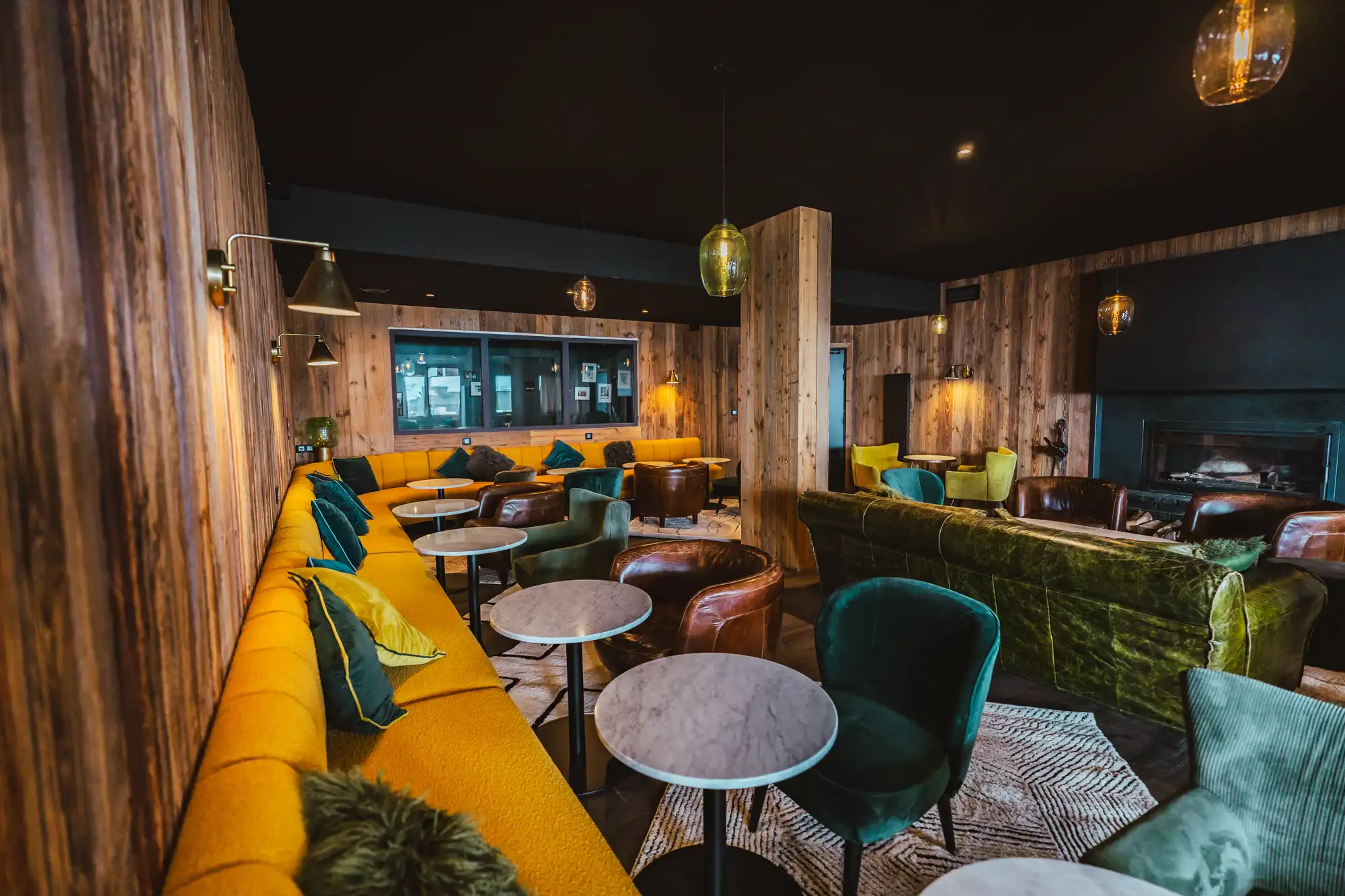 Salon moderne avec murs en bois, grand canapé jaune, fauteuils verts et marron, tables rondes et cheminées.