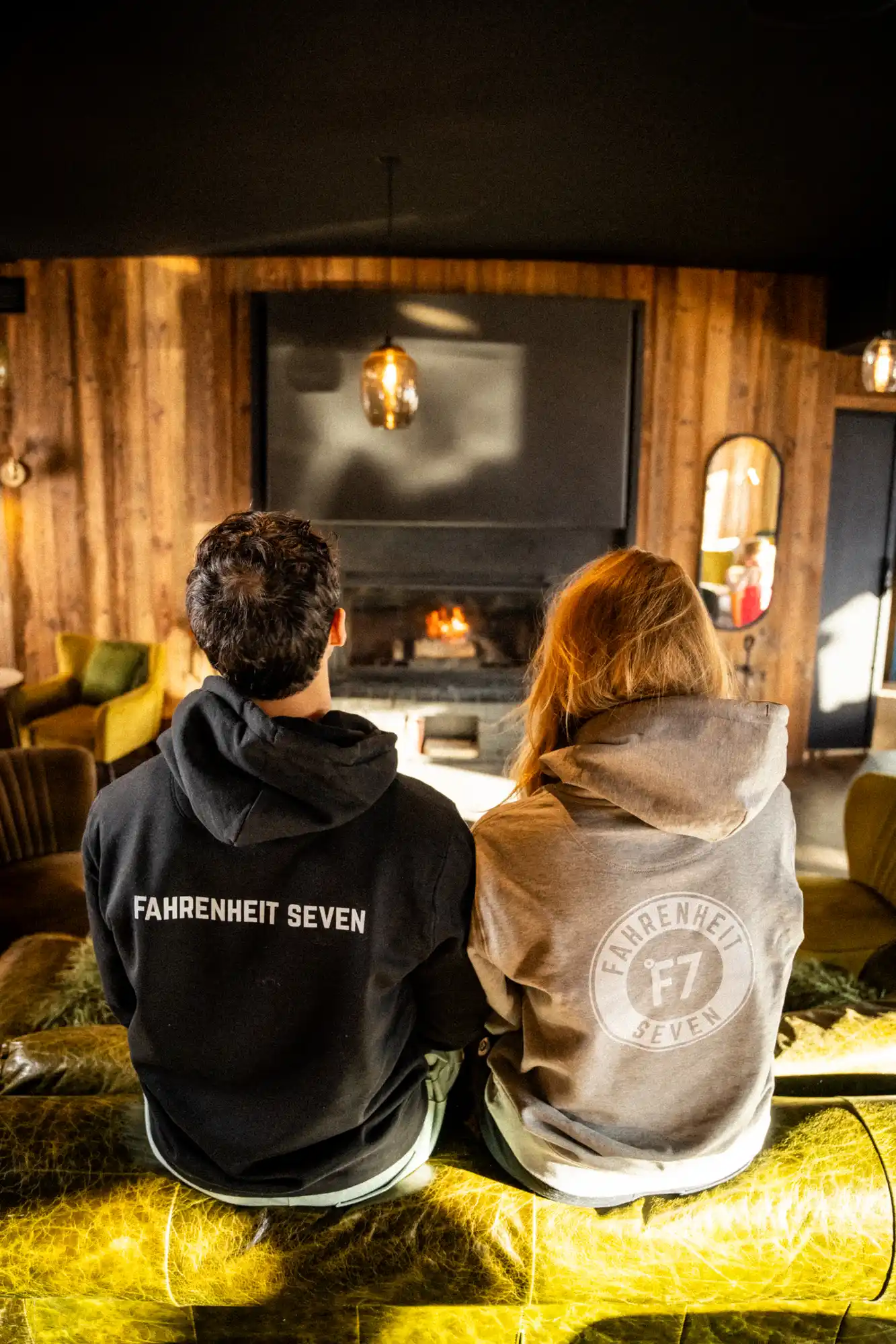 Deux personnes vues de dos, assises sur un canapé vert olive dans un salon au mur en bois, portant des sweat-shirts avec le texte « Fahrenheit Seven » et un logo « F7 » sur le dos, devant une cheminée allumée.