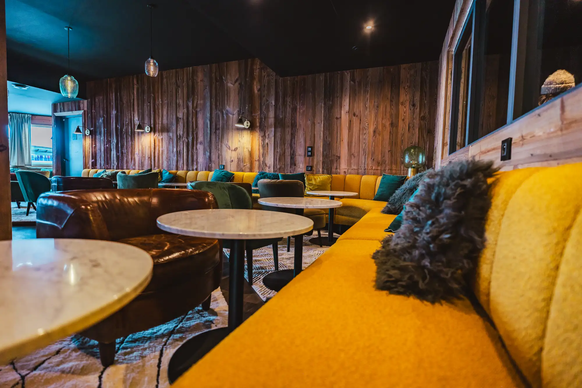 Intérieur d'un salon confortable avec un canapé d'angle jaune, coussins verts et gris, fauteuil en cuir marron, tables basses rondes à plateaux blancs, murs en bois foncé, et éclairage tamisé.