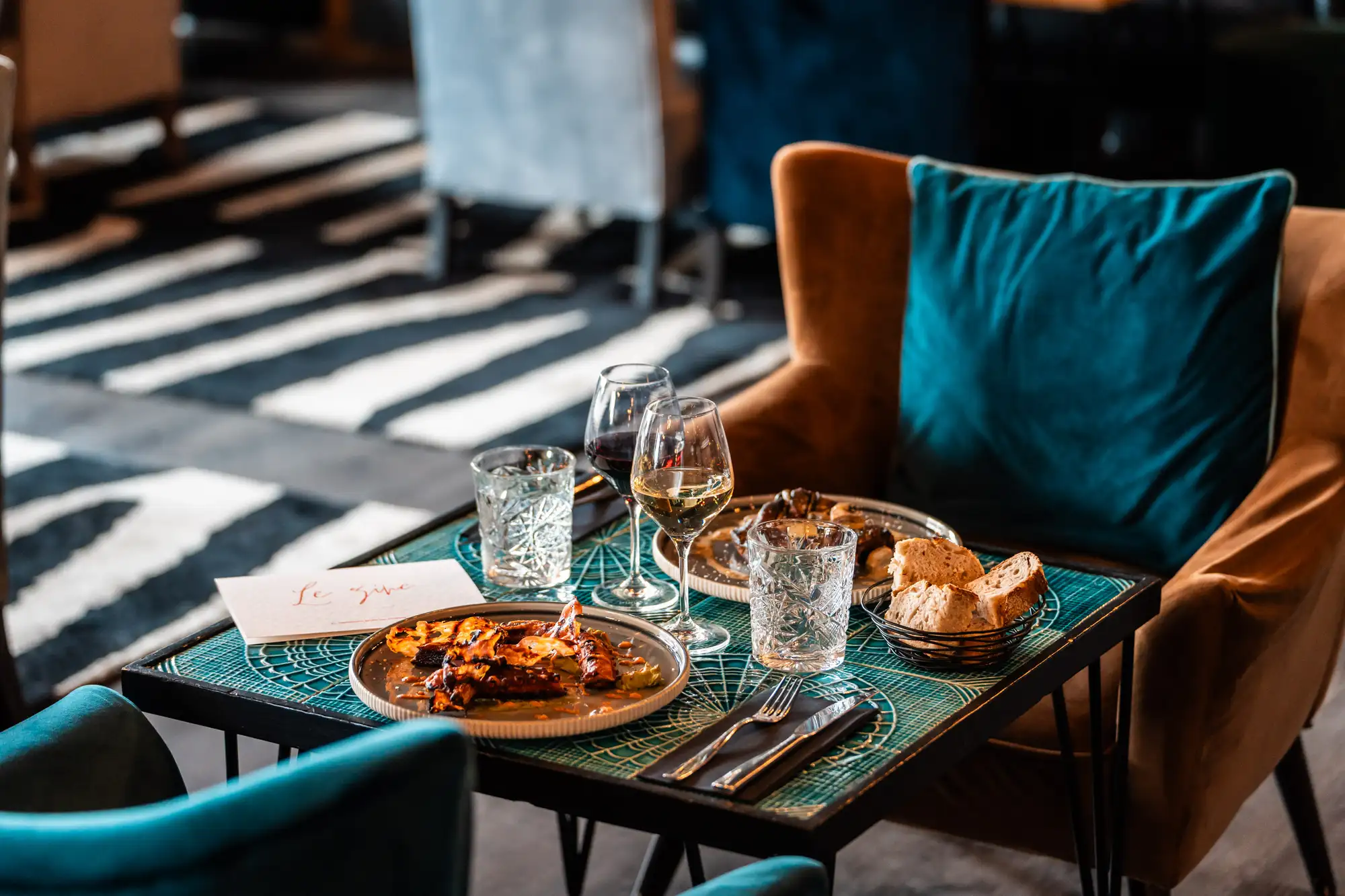 Table de restaurant avec chaise bleu et marron, servie avec deux assiettes de plats gourmets, deux verres de vin rouge et blanc, deux verres d'eau, un panier de pain et une carte restaurant posée.