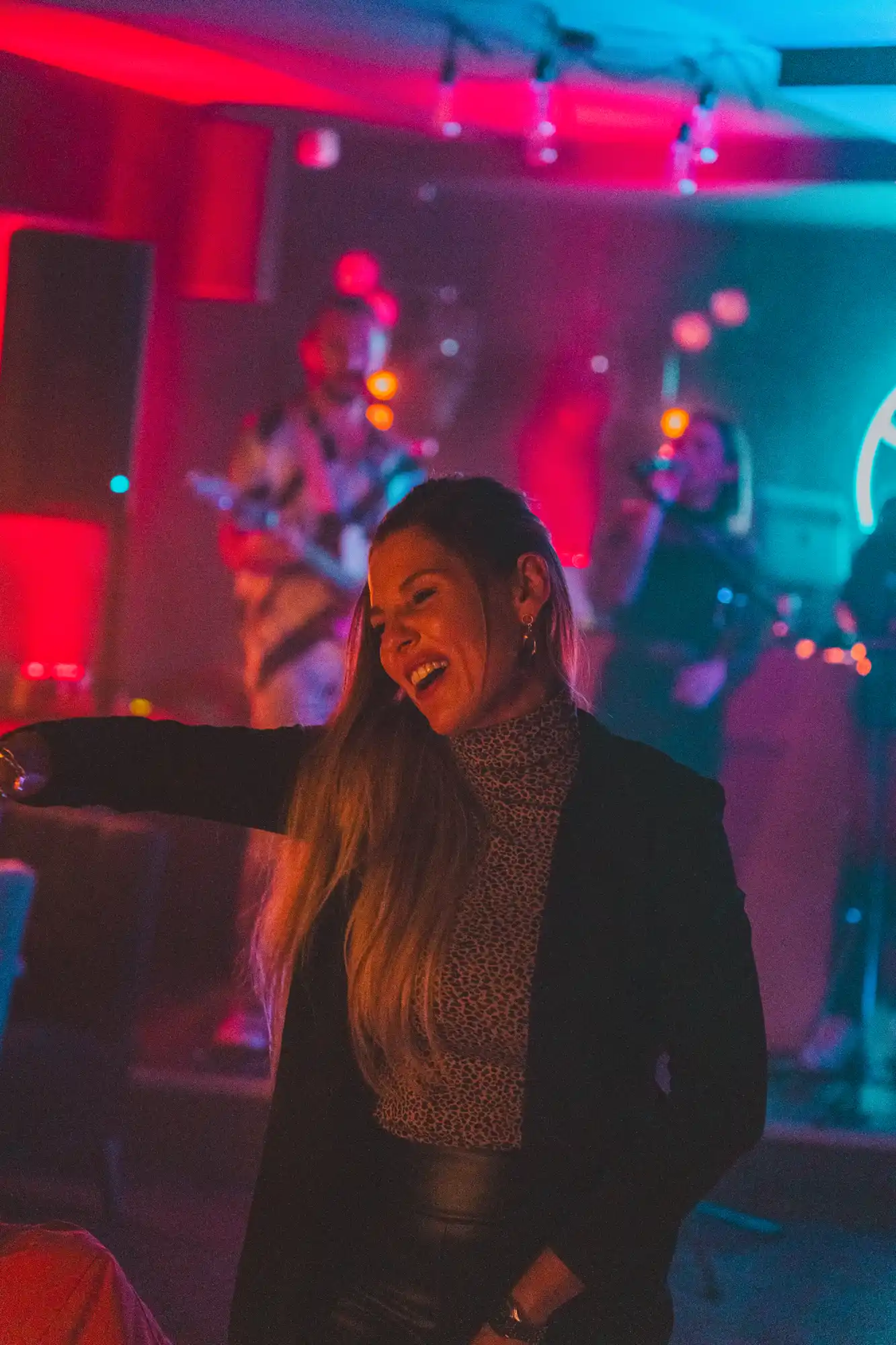Une femme souriante aux cheveux longs danse dans un club avec un éclairage rouge et bleu, tandis qu'un groupe de musiciens joue en arrière-plan.