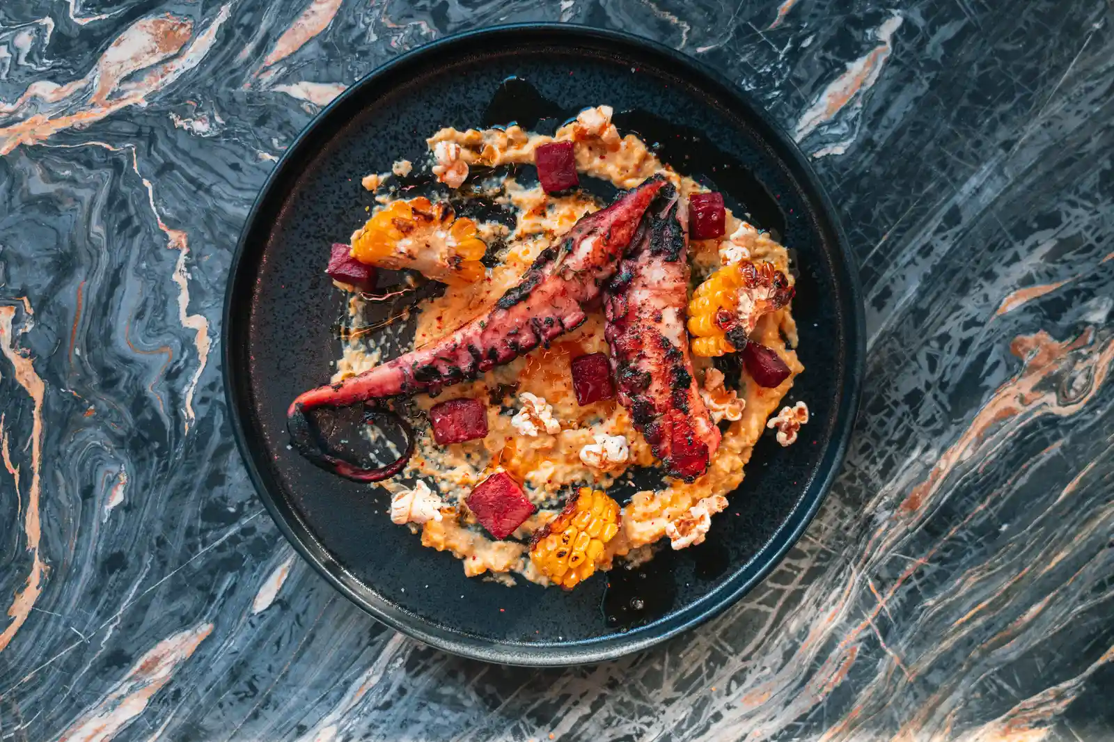 Plat noir contenant deux tentacules de pieuvre grillées sur une purée orange, avec morceaux de maïs grillé, cubes rouges et éclats de pop-corn, le tout présenté sur un fond de table bleu-gris et blanc veinée.