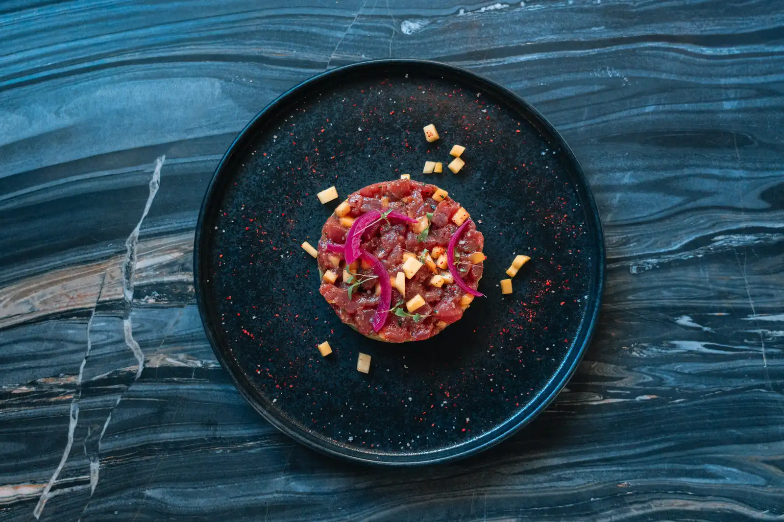 Tartare de poisson rouge présenté en cercle sur une assiette noire avec des petits dés jaunes, des oignons rouges en rondelles, et des herbes, sur une table en marbre à motifs bleus.