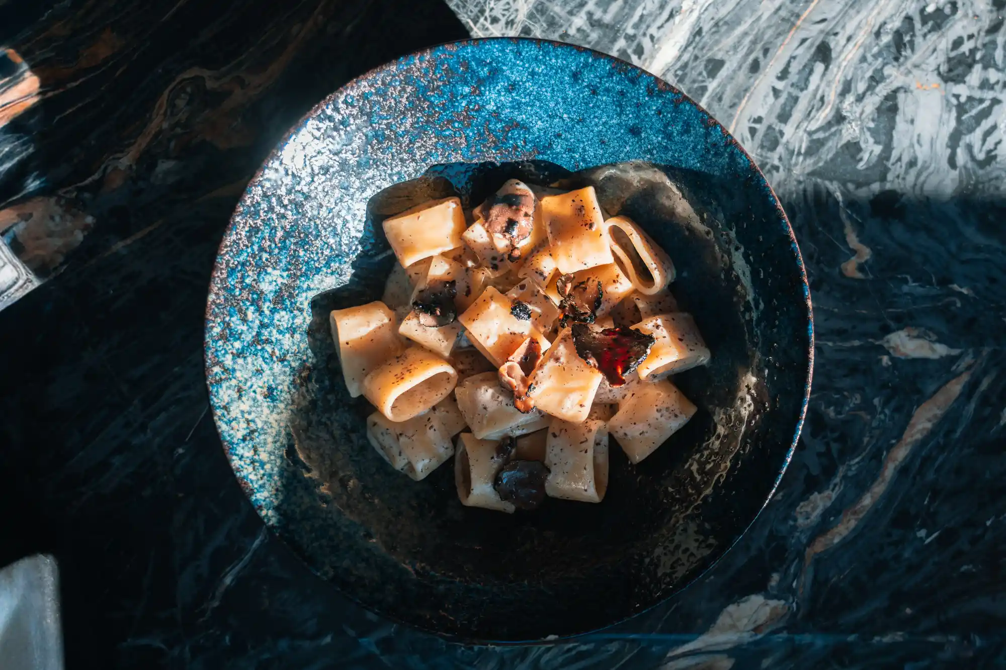 Plat de pâtes rigatoni dans une sauce crémeuse, garnies de morceaux de truffe, servies dans une assiette bleue sur une table en marbre sombre.