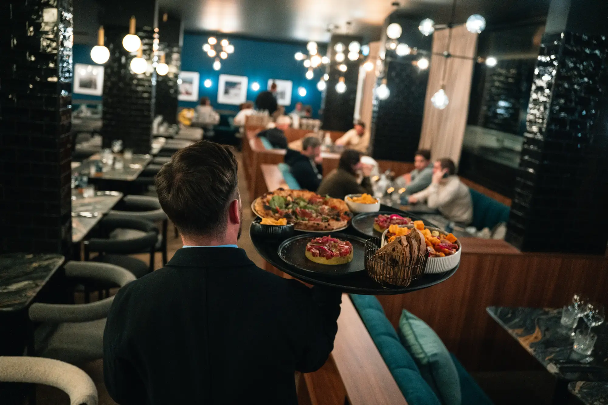 Serveur portant un plateau avec pizzas, pain et garnitures dans un restaurant moderne éclairé avec des clients assis en arrière-plan.