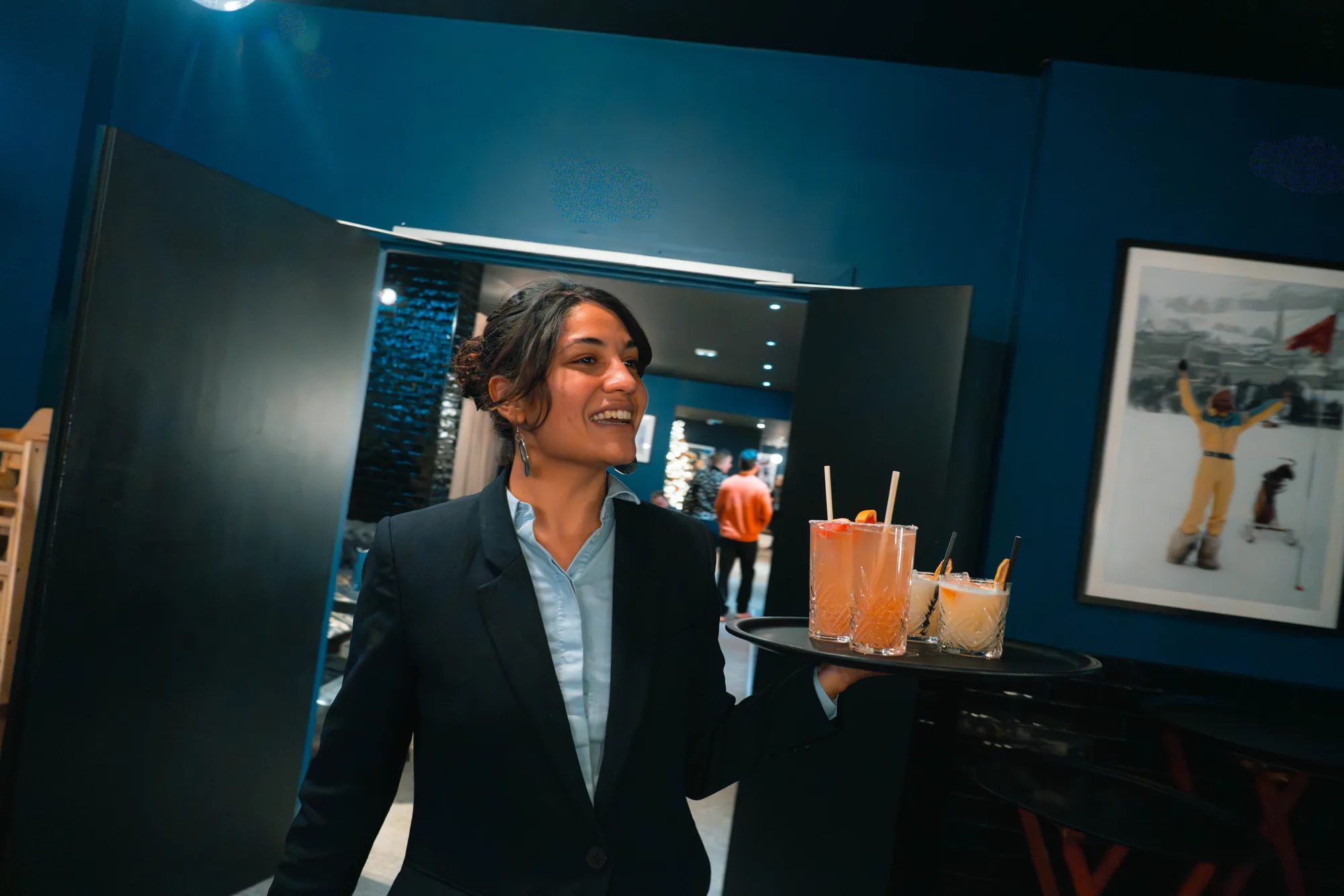 Serveuse souriante portant un plateau avec quatre cocktails colorés dans un restaurant au décor moderne bleu et noir.