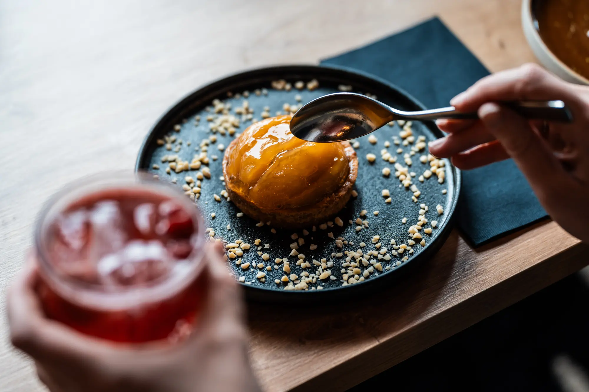 Une main tient une cuillère proche d'une petite tarte avec un dessus brillant et orangé sur une assiette noire parsemée de morceaux de noix, avec une autre main tenant un verre de boisson rouge en arrière-plan.