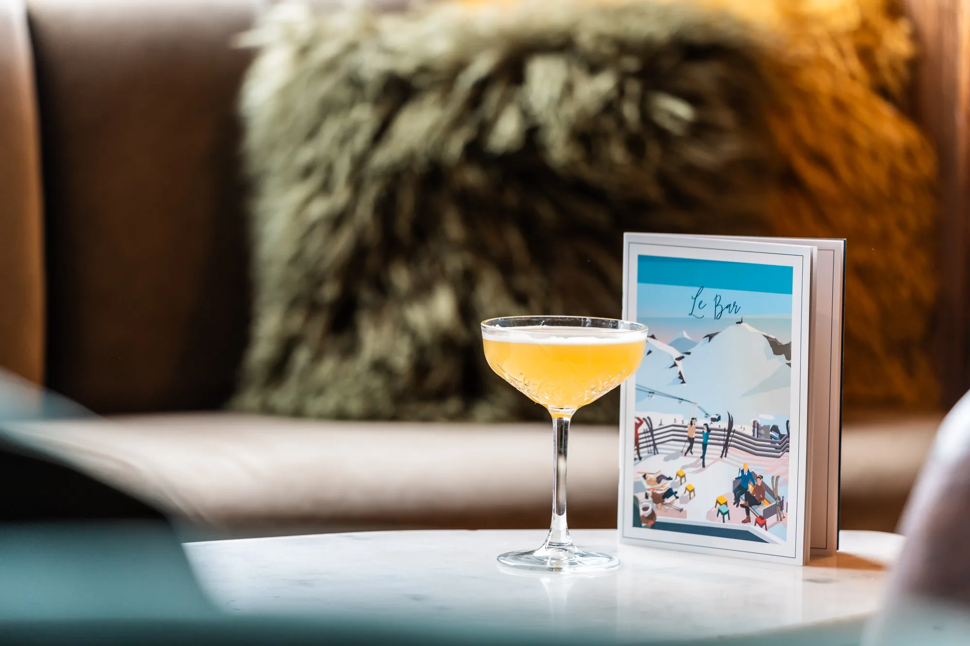 Un verre de cocktail orange clair sur une table blanche avec un menu montrant une scène de montagne enneigée et le titre « Le Bar », avec un canapé marron et un coussin pelucheux en arrière-plan flou.