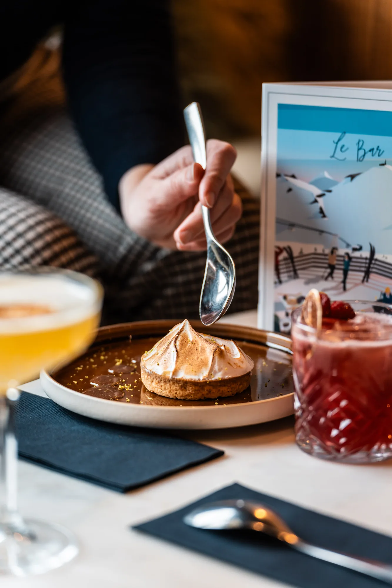 Main tenant une cuillère au-dessus d'une tartelette avec un dessus meringué doré sur une assiette, avec deux verres de boissons sur une table et un menu en arrière-plan.