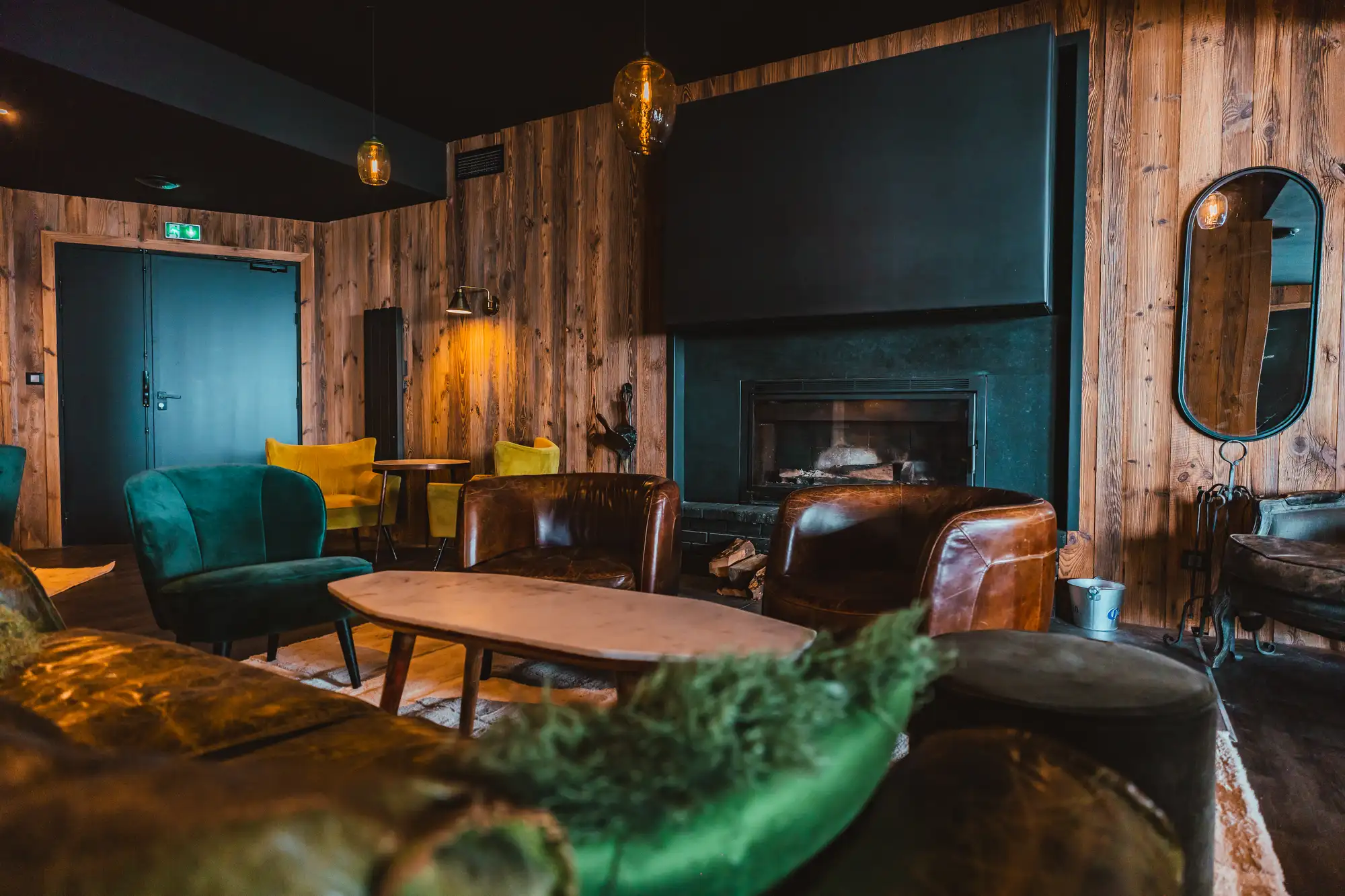 Salon chaleureux avec murs en bois, cheminée noire, fauteuils en cuir marron et velours vert, chaises jaunes, grande porte bleu foncé et miroir ovale.