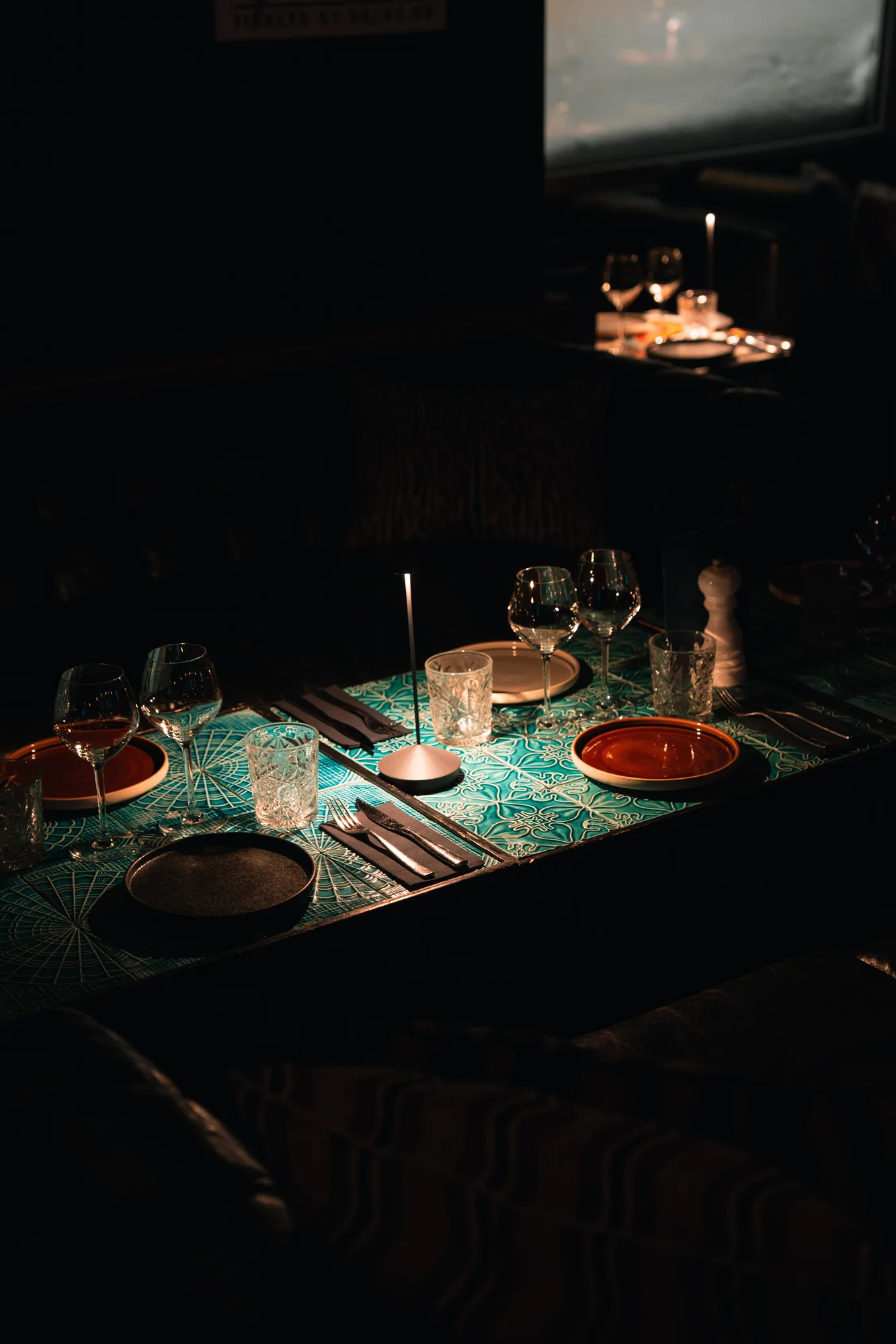 Table sombrement éclairée avec nappe à motifs verts, verres à vin et à eau, assiettes rouges et noires, couverts et lampe fine au centre.