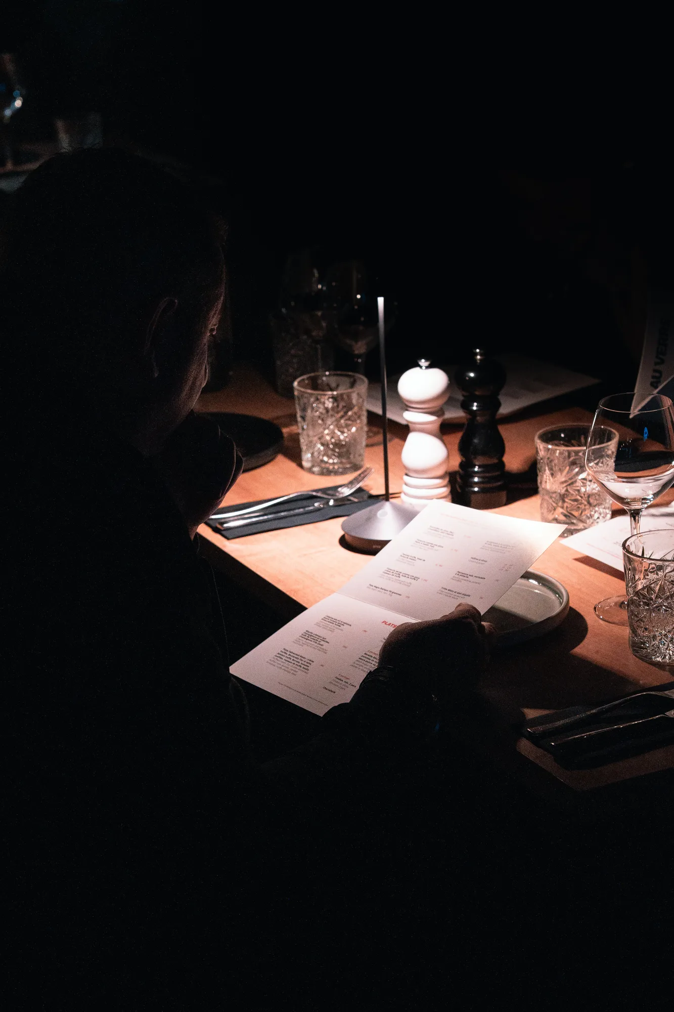 Une personne assise à une table faiblement éclairée dans un restaurant, tenant un menu cartonné, entourée de verres, d'un moulin à sel blanc, d'un moulin à poivre noir, de couverts, et d'une petite lampe.