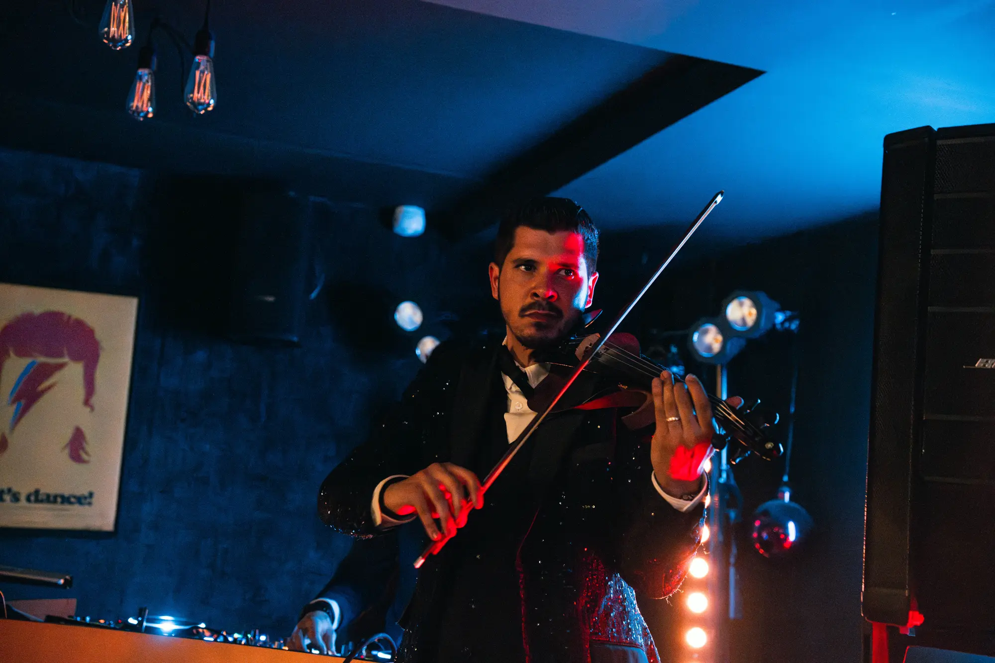 Un homme joue du violon électrique sur scène sous un éclairage bleu et rouge, portant un costume noir brillant et une chemise blanche.
