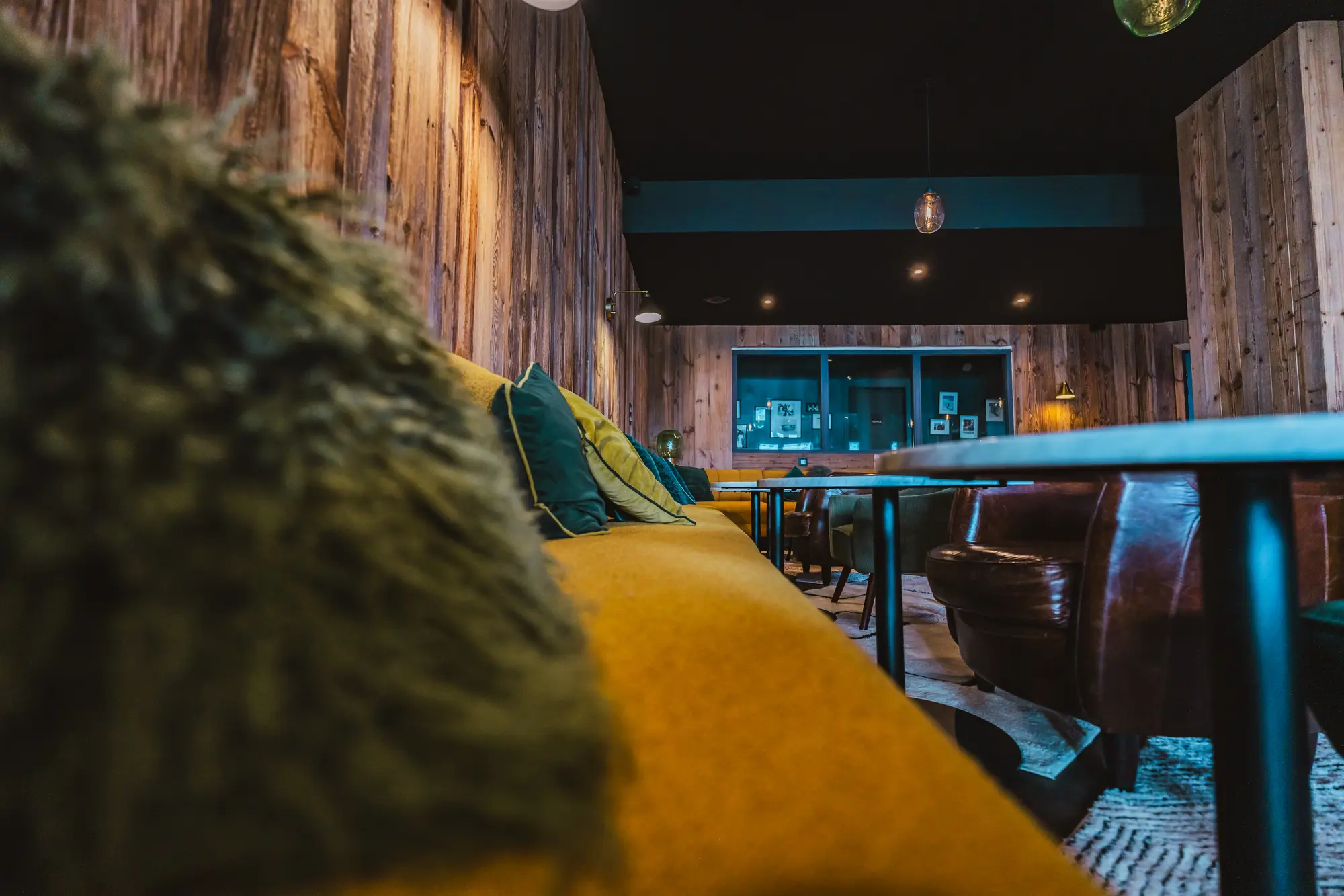 Intérieur d'un salon ou café avec un canapé jaune à coussins verts et bleus, des murs en bois, plusieurs fauteuils en cuir brun et des petites tables noires sur un tapis à motifs.