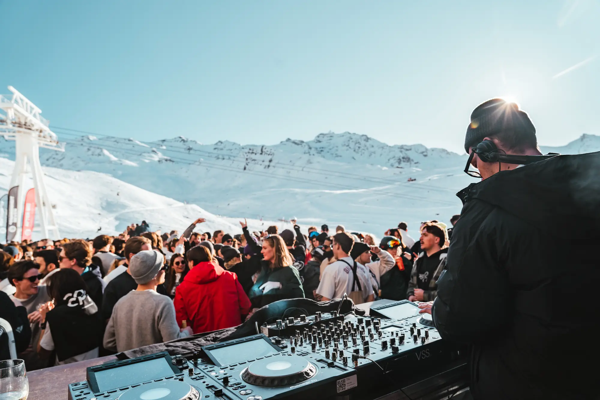 Un DJ portant un bonnet noir et des lunettes joue de la musique sur une console DJ devant une foule en plein air dans un paysage enneigé de montagne sous un ciel bleu clair.