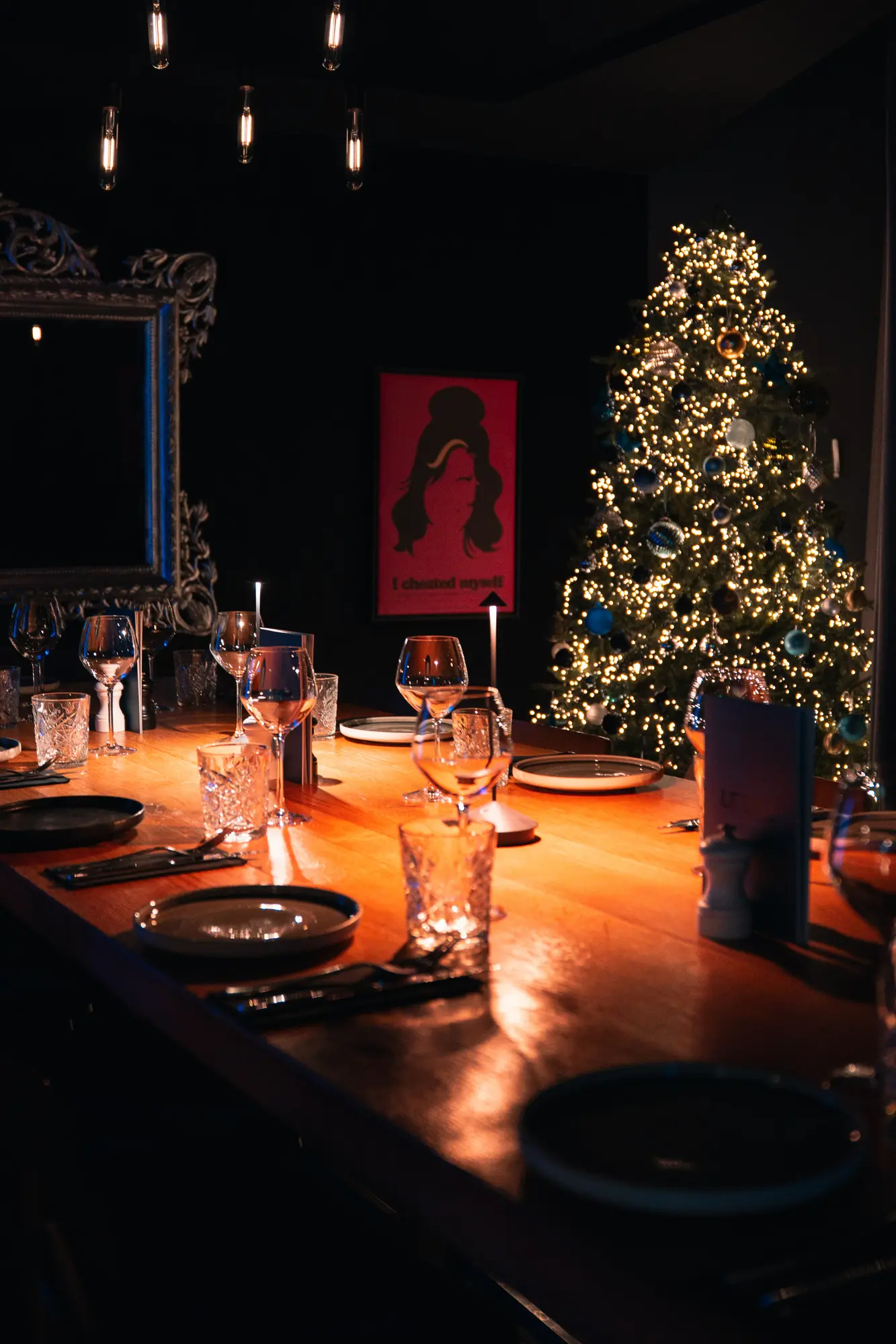 Table en bois avec des verres, assiettes et couverts dressée pour un repas, éclairée par des lumières chaudes, avec un sapin de Noël décoré et illuminé en arrière-plan dans une pièce sombre.