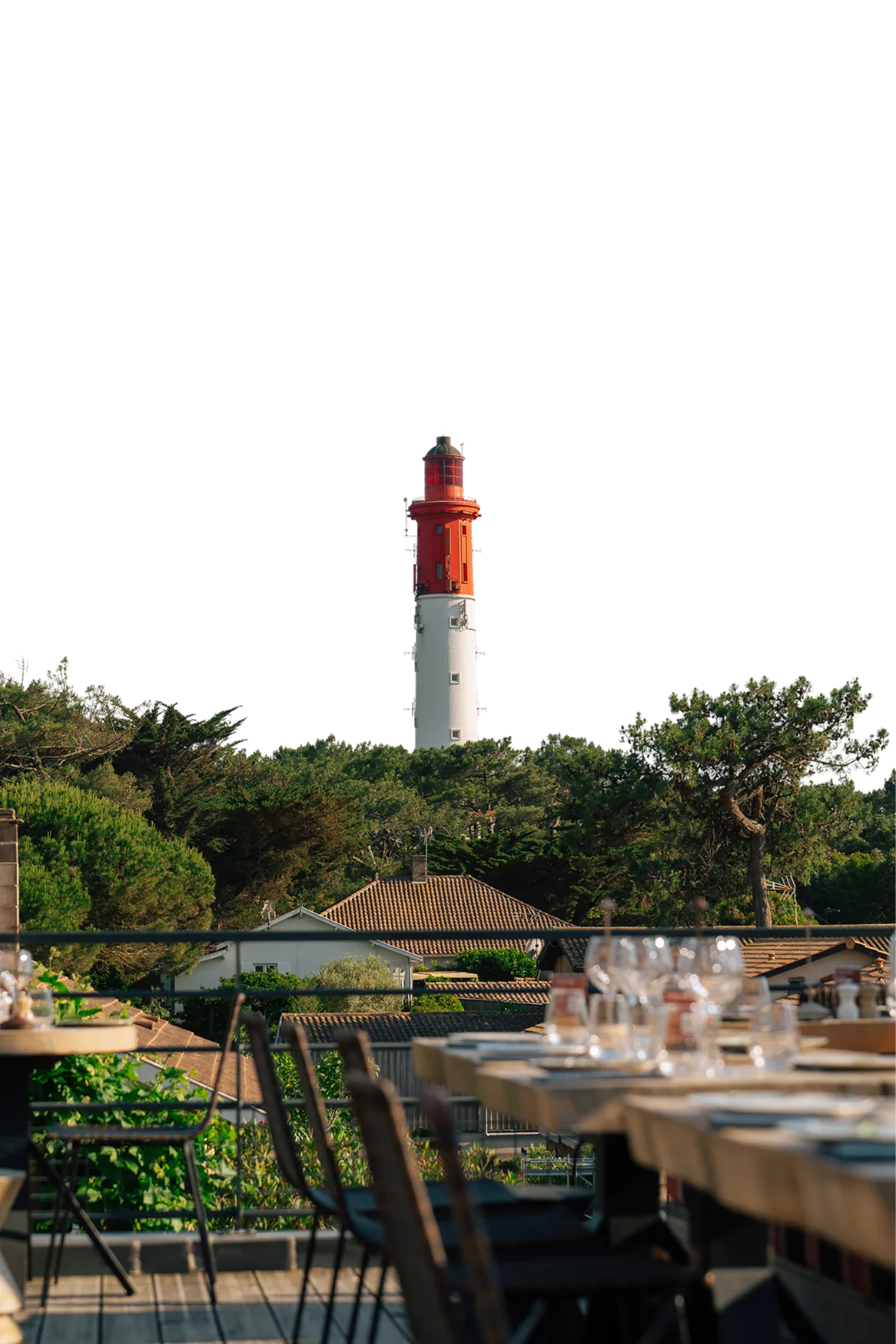 Phare rouge et blanc se dressant derrière des arbres verts et des toits de maisons, vu depuis une terrasse avec des tables et des chaises.