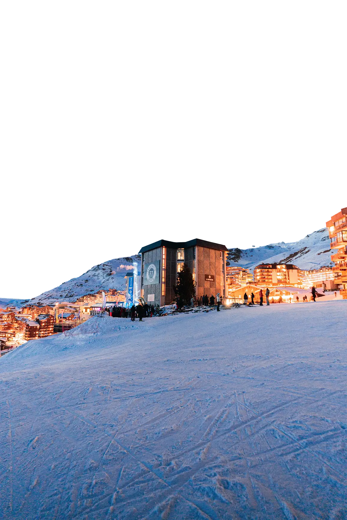 Station de ski en soirée avec un bâtiment central éclairé et des skieurs sur la neige, entourée de résidences en montagne enneigée.