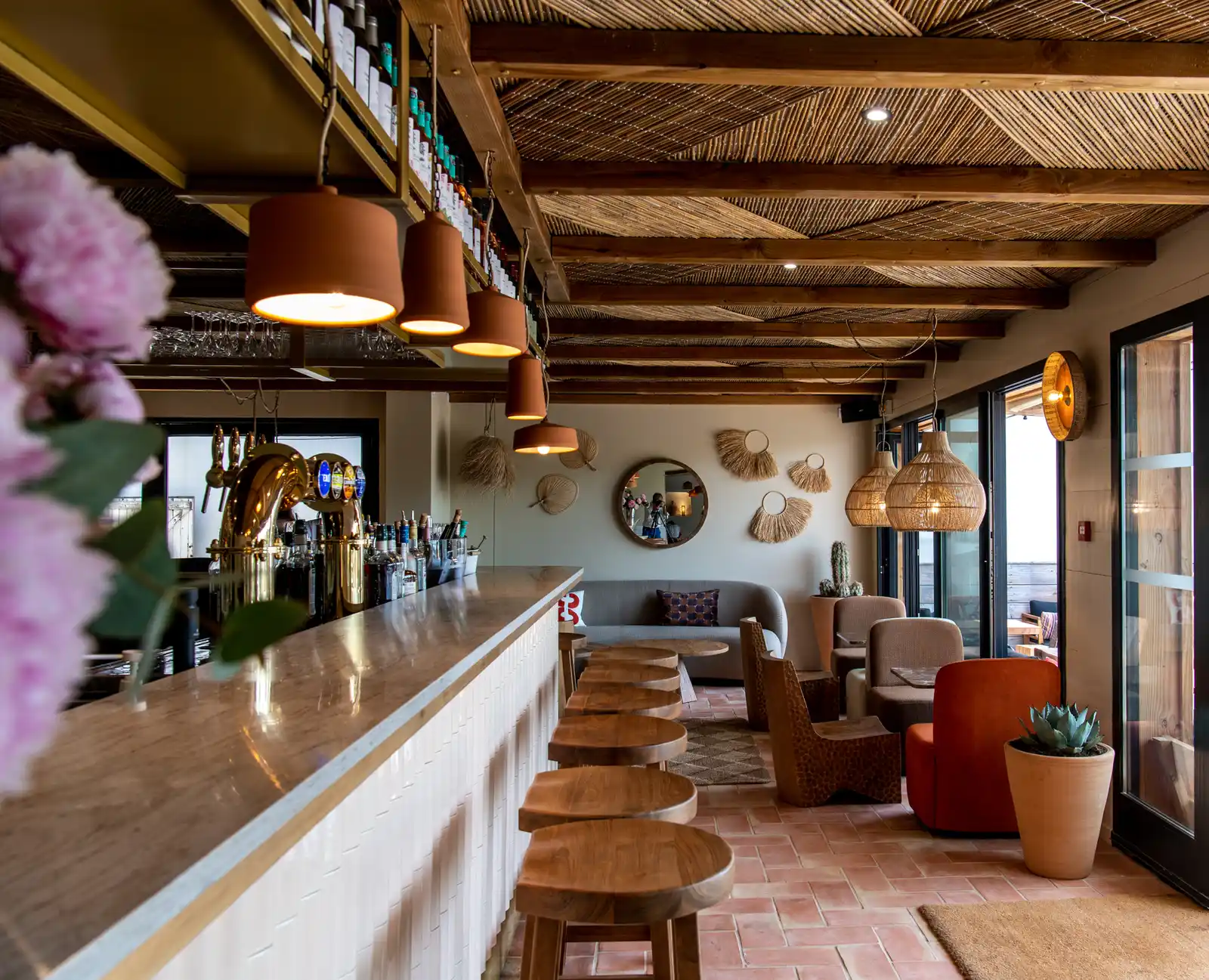 Intérieur d'un bar moderne avec un comptoir en marbre et des tabourets en bois alignés, éclairage suspendu en terre cuite, murs décorés de miroirs ronds et d'éléments en fibres naturelles, sièges confortables en tissu et plantes en pot près des grandes fenêtres vitrées.