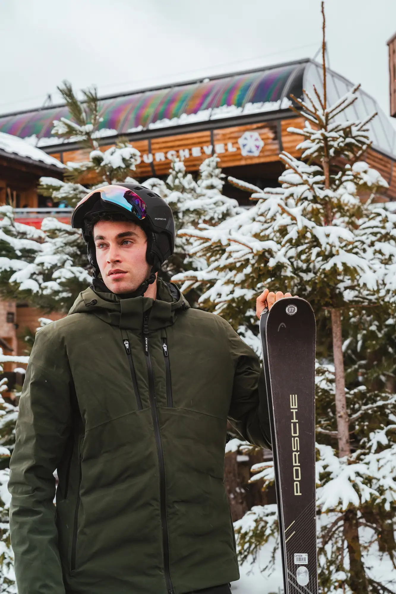 Un homme en veste verte, casque noir avec visière réfléchissante, tient un ski noir marqué Porsche devant des sapins enneigés et un bâtiment en bois avec une enseigne indiquant Courchevel.