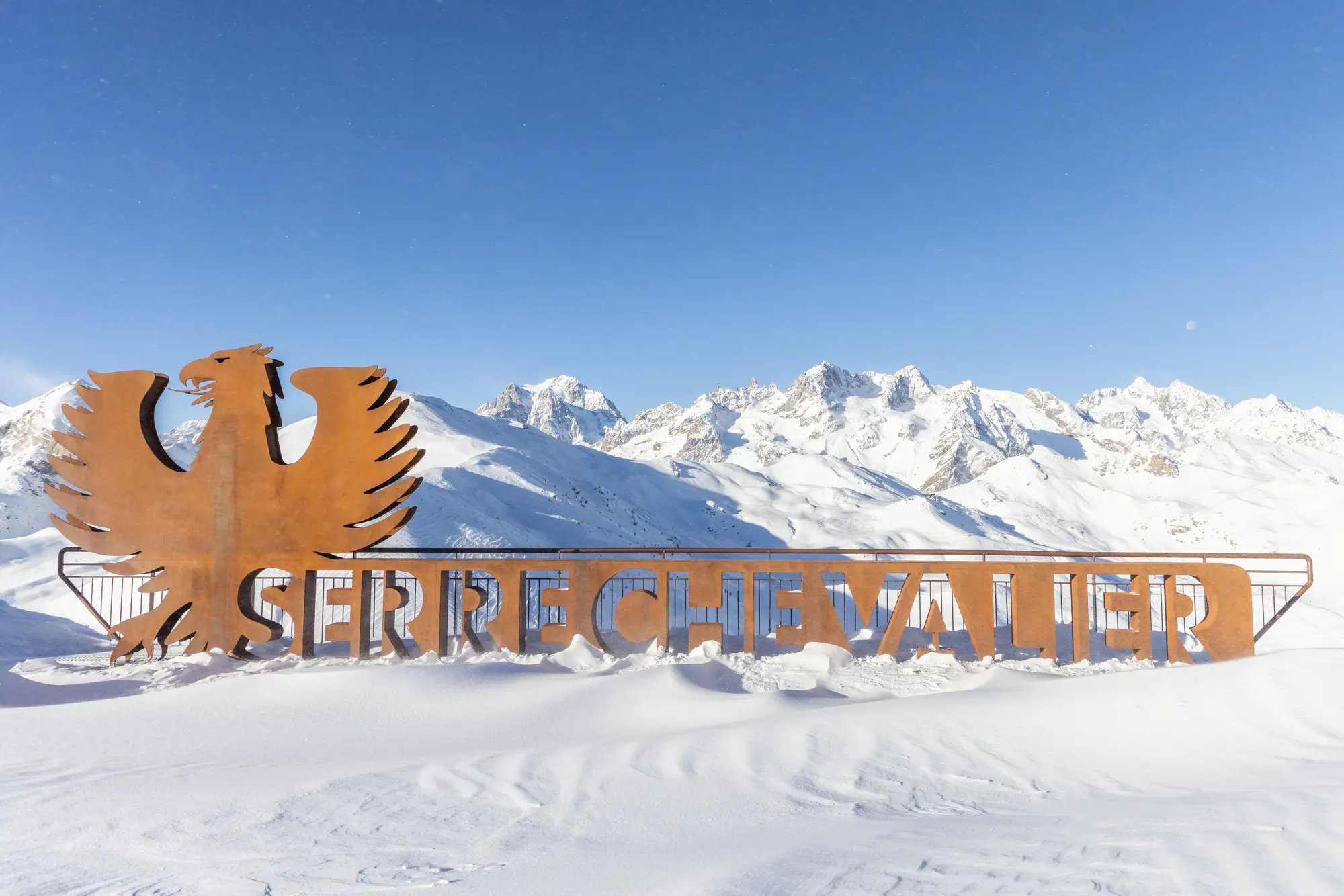 Sculpture en métal rouillé représentant un aigle stylisé et les lettres formant le nom 'Serre Chevalier' installée dans un paysage enneigé avec une chaîne de montagnes en arrière-plan sous un ciel bleu clair.