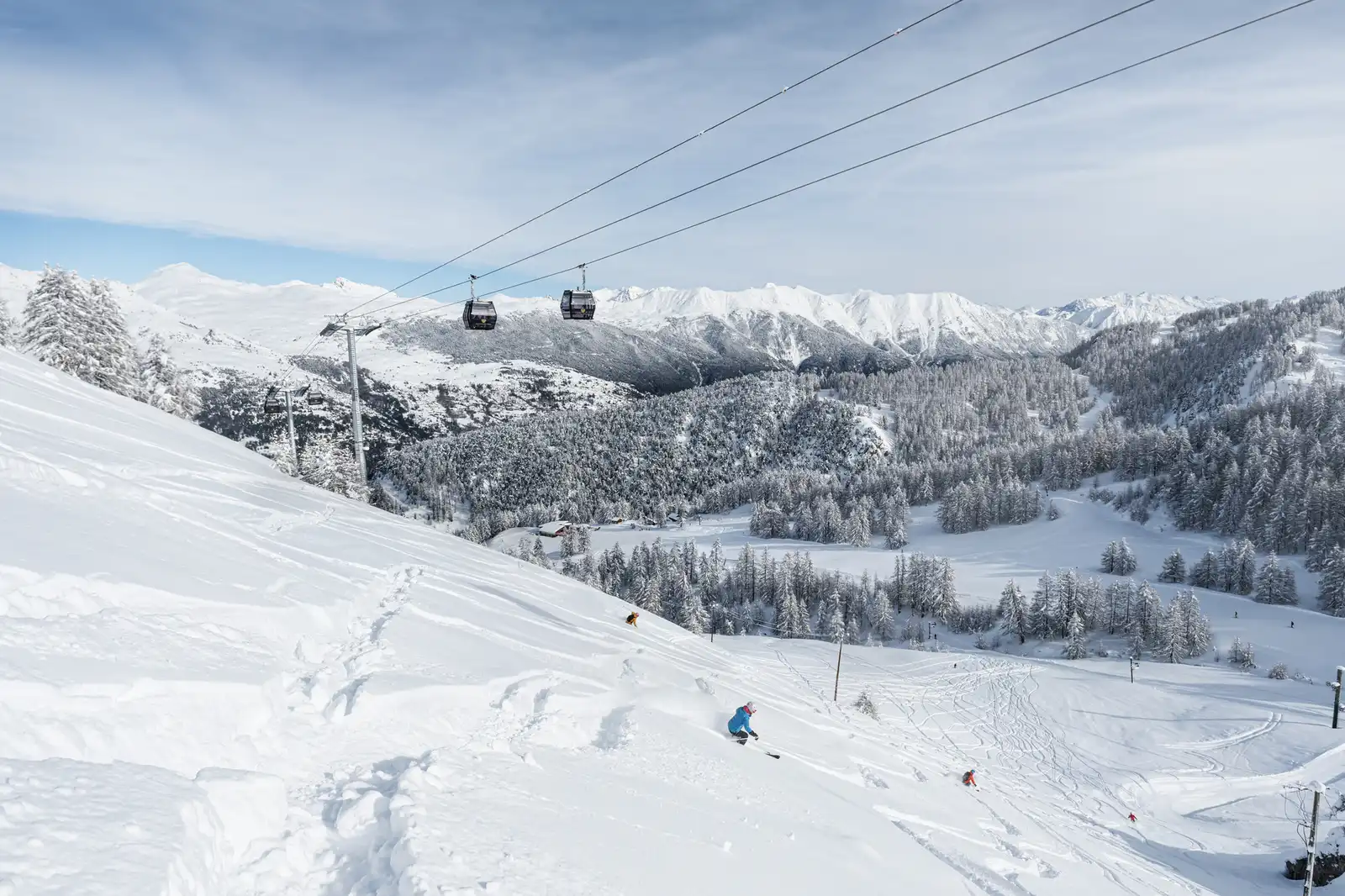 Station de ski avec des skieurs descendant une pente enneigée, des télécabines suspendues au-dessus, et une vaste forêt de sapins enneigés avec des montagnes en arrière-plan sous un ciel clair.