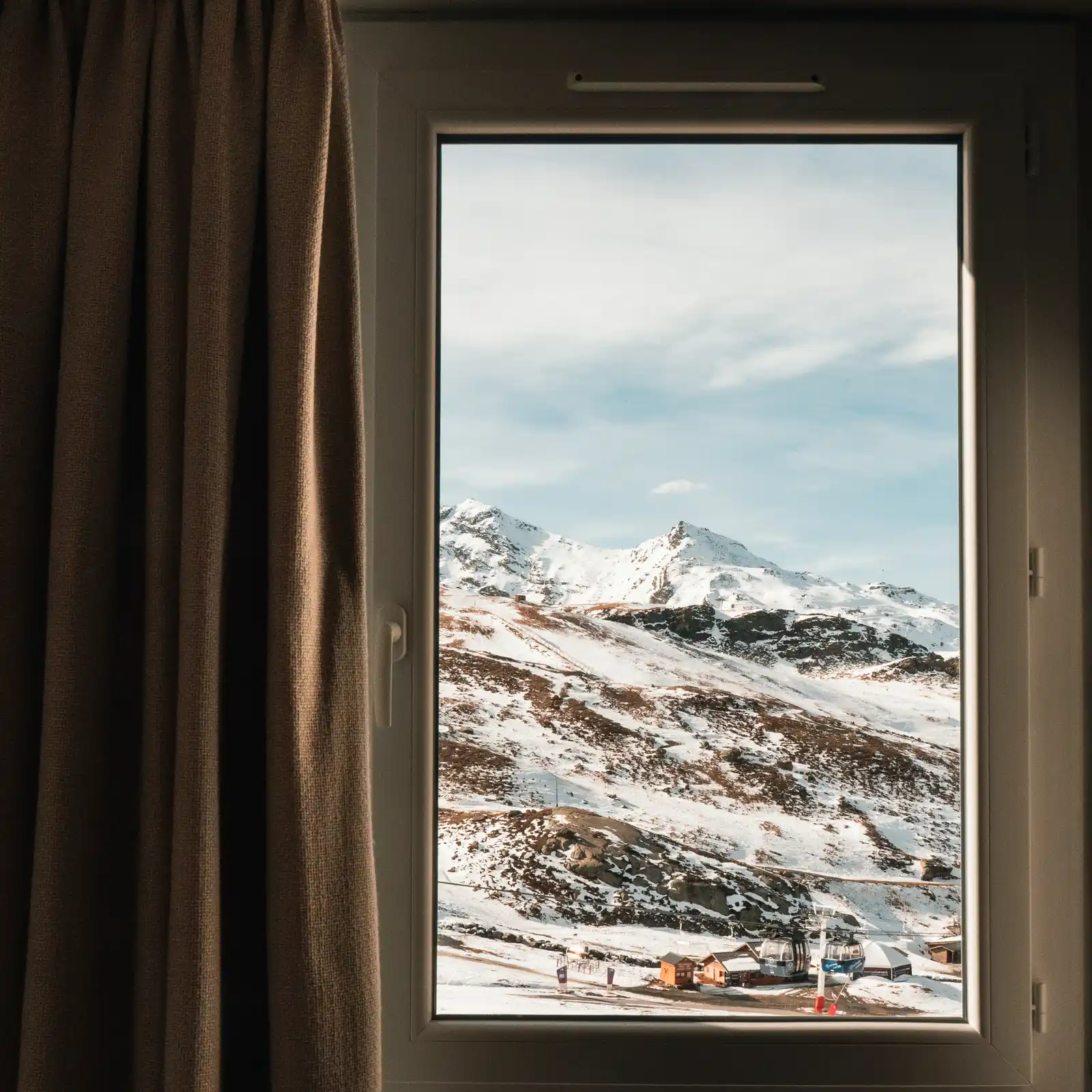 Vue par une fenêtre d'un paysage de montagne enneigée avec un ciel partiellement nuageux, des télécabines et des chalets au premier plan.