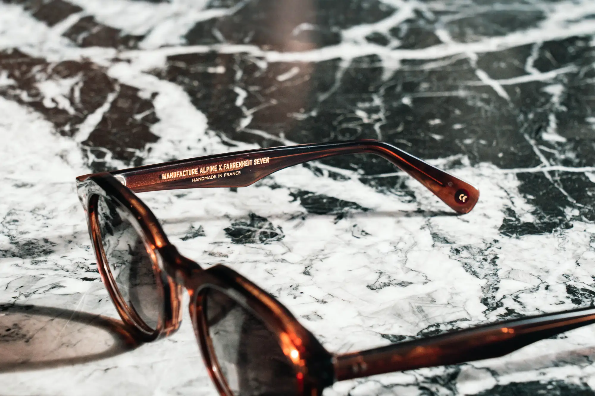 Lunettes de soleil marron transparent posées sur une surface en marbre noir et blanc, avec l'inscription sur la branche indiquant "Manufacture Alpine x Fahrenheit Seven, fait main en France".