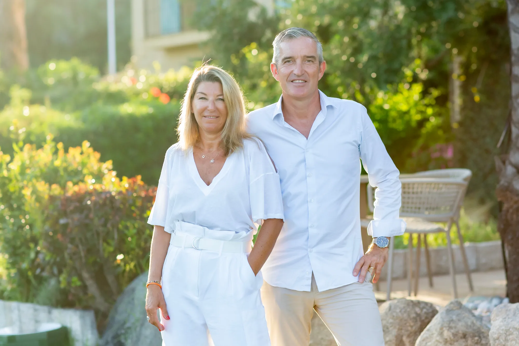 Un homme et une femme souriants debout côte à côte dans un jardin ensoleillé, tous deux vêtus de vêtements blancs décontractés.