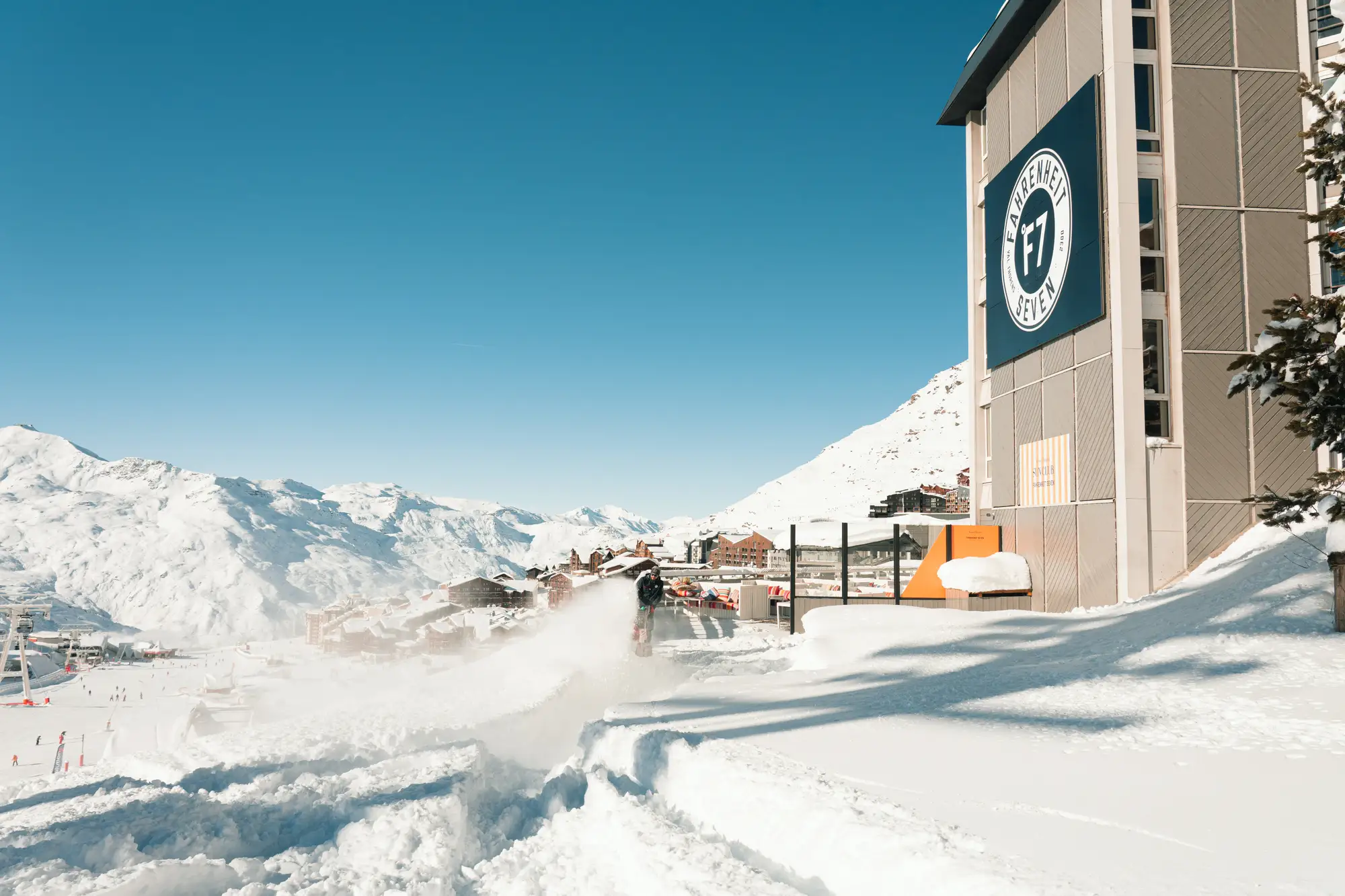Un homme nettoie la neige sur un chemin à côté d'un bâtiment avec un panneau Fahrenheit Seven dans une station de ski entourée de montagnes enneigées sous un ciel bleu clair.